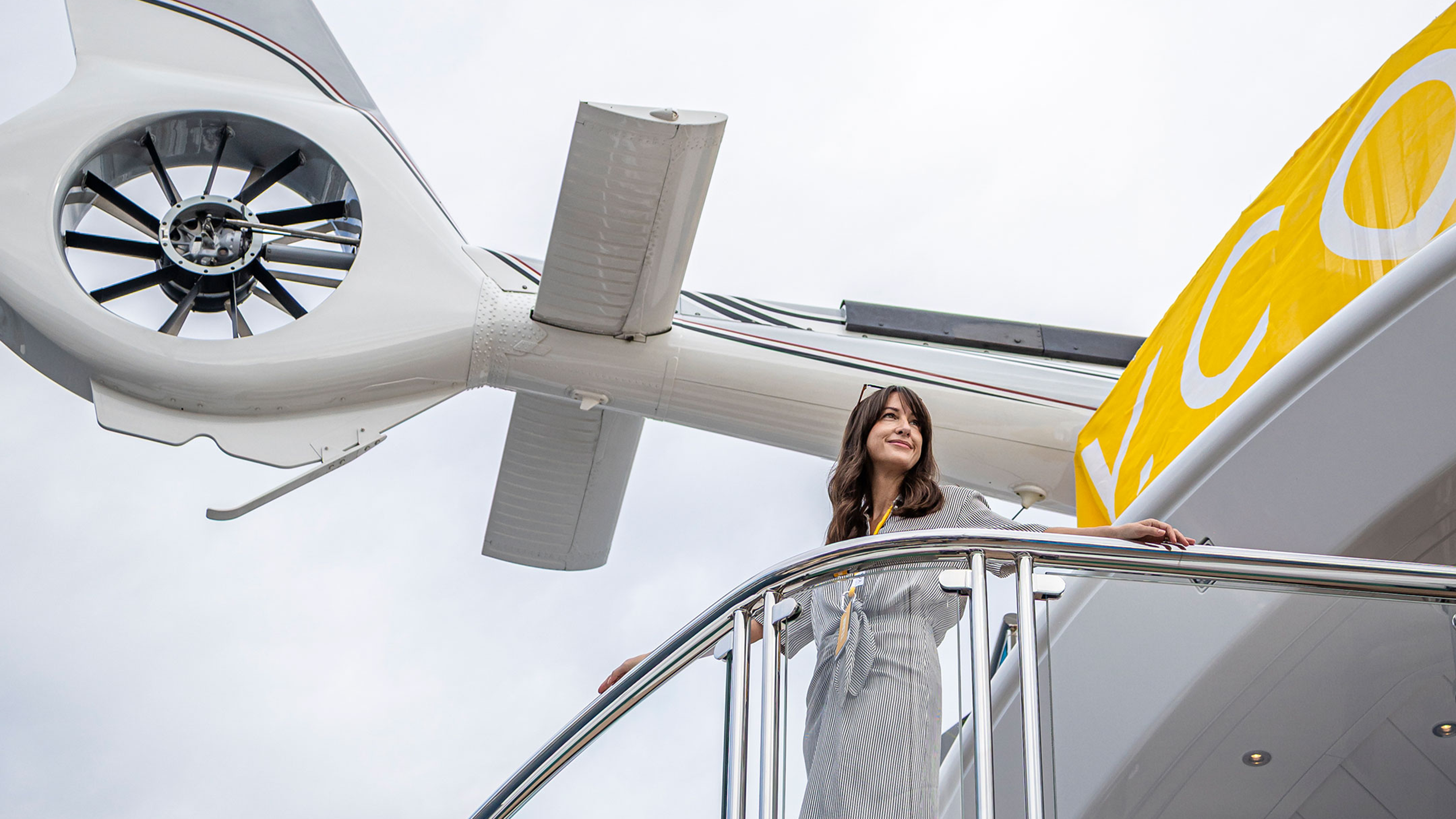 A woman in a grey dress stands on a balcony in the foreground, with a large white aircraft and a yellow banner in the background.