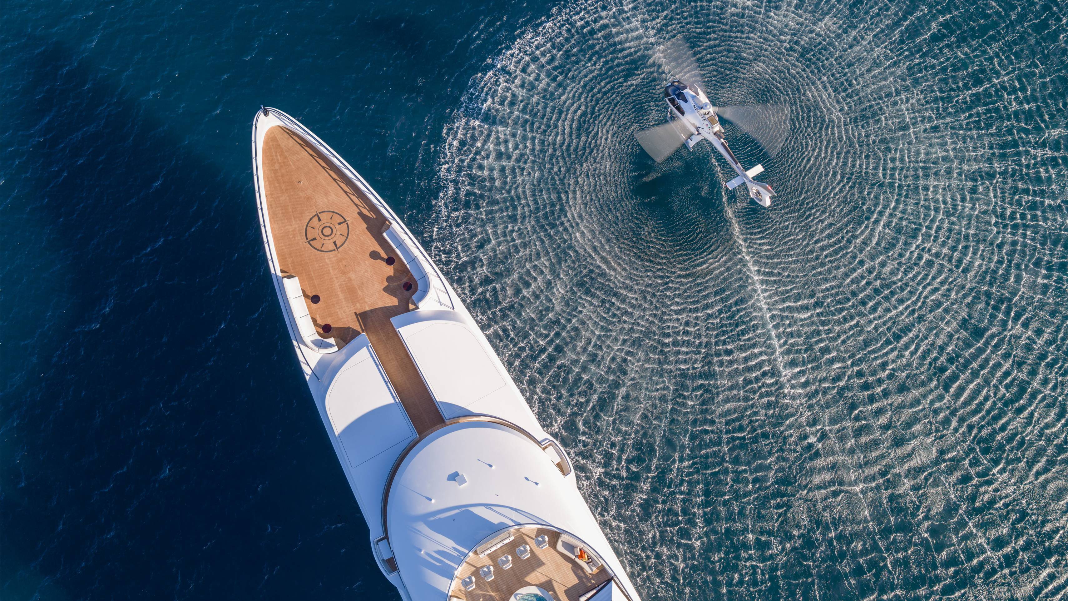A helicopter creates concentric ripples on the deep blue sea beside a luxurious white yacht.