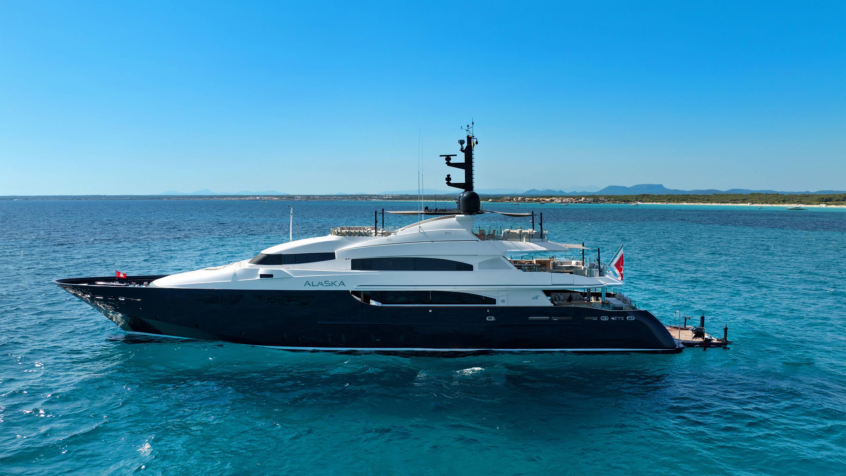 A large, dark-hulled yacht named "ALASKA" floats on turquoise water in the foreground, with a distant, hazy coastline under a clear blue sky in the background.