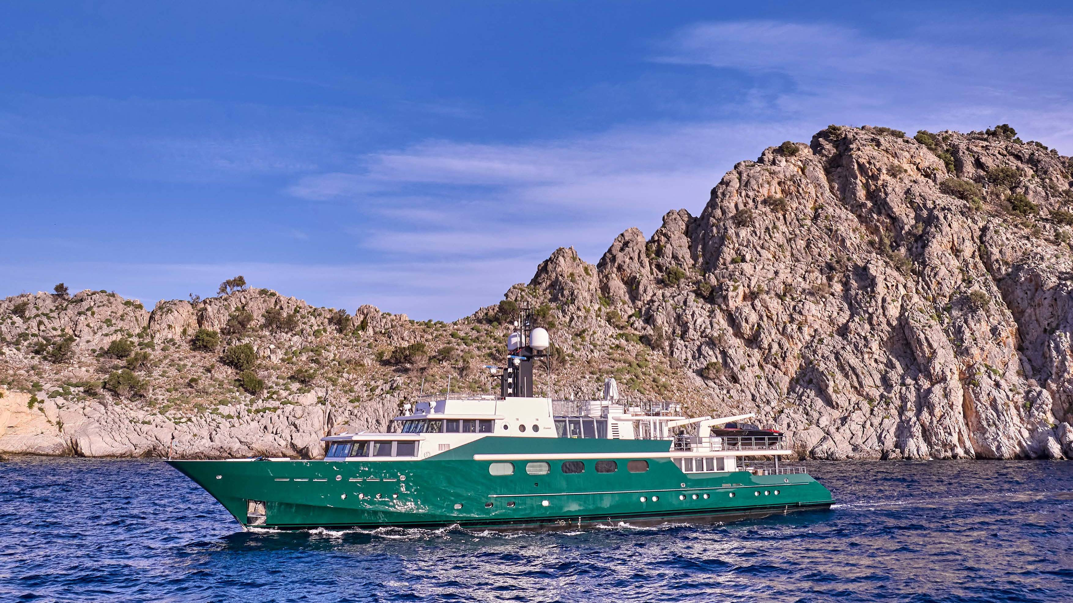 A green yacht sails on blue water in the foreground, with rugged, rocky cliffs and a clear blue sky in the background.