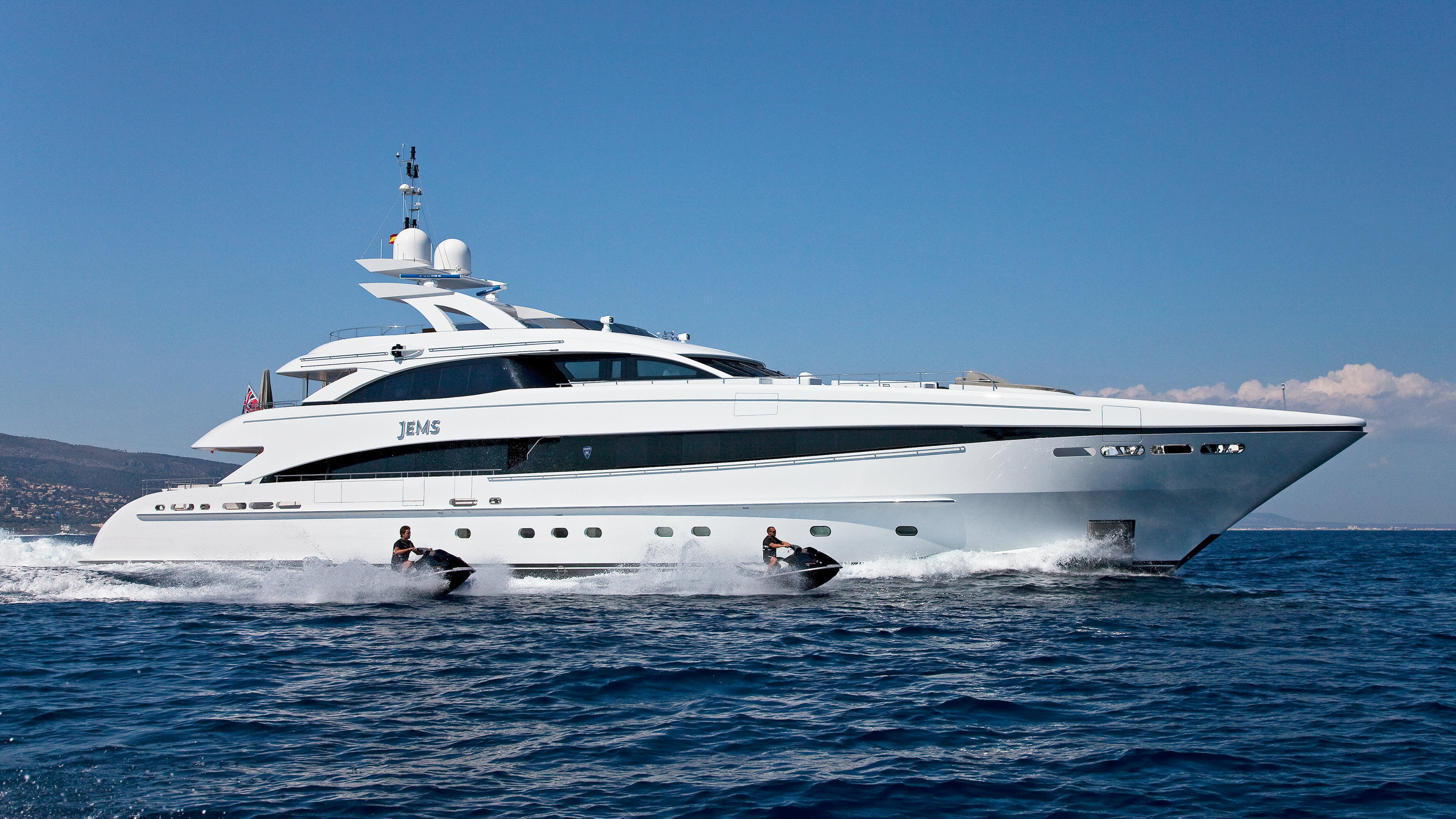 A sleek white yacht cruises through the deep blue ocean, trailed by two jet skiers creating a foamy wake.