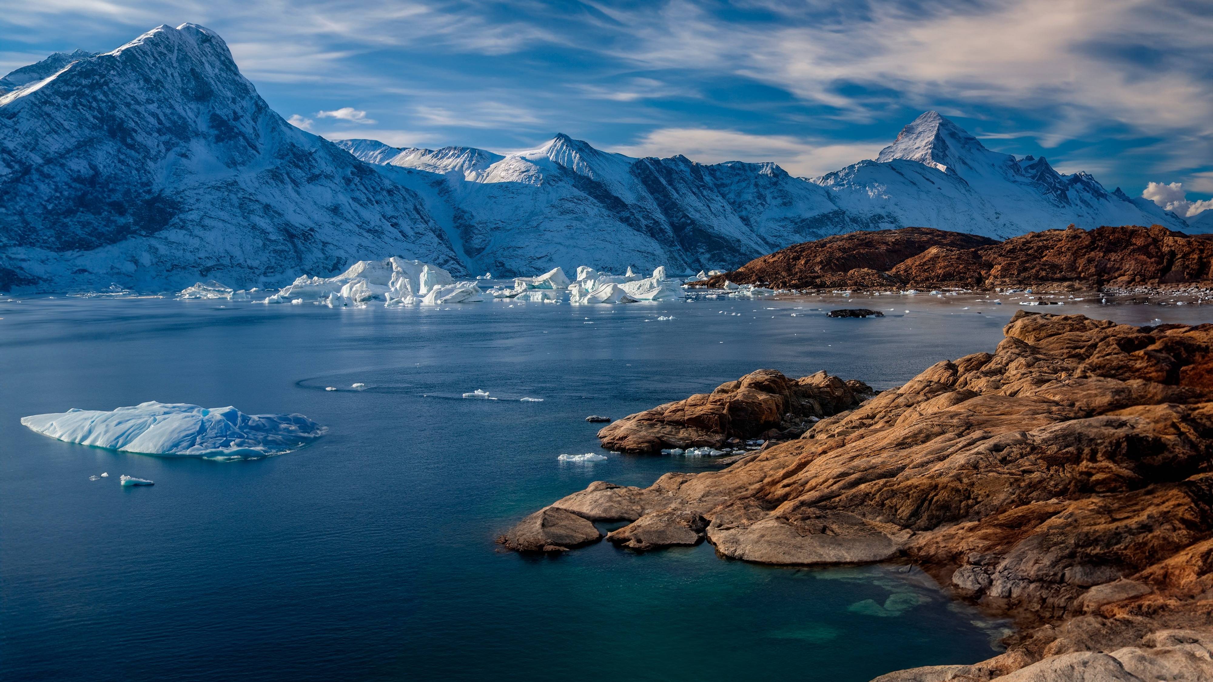 Majestic snow-capped mountains rise above a serene bay dotted with icebergs and rugged, rocky shores.