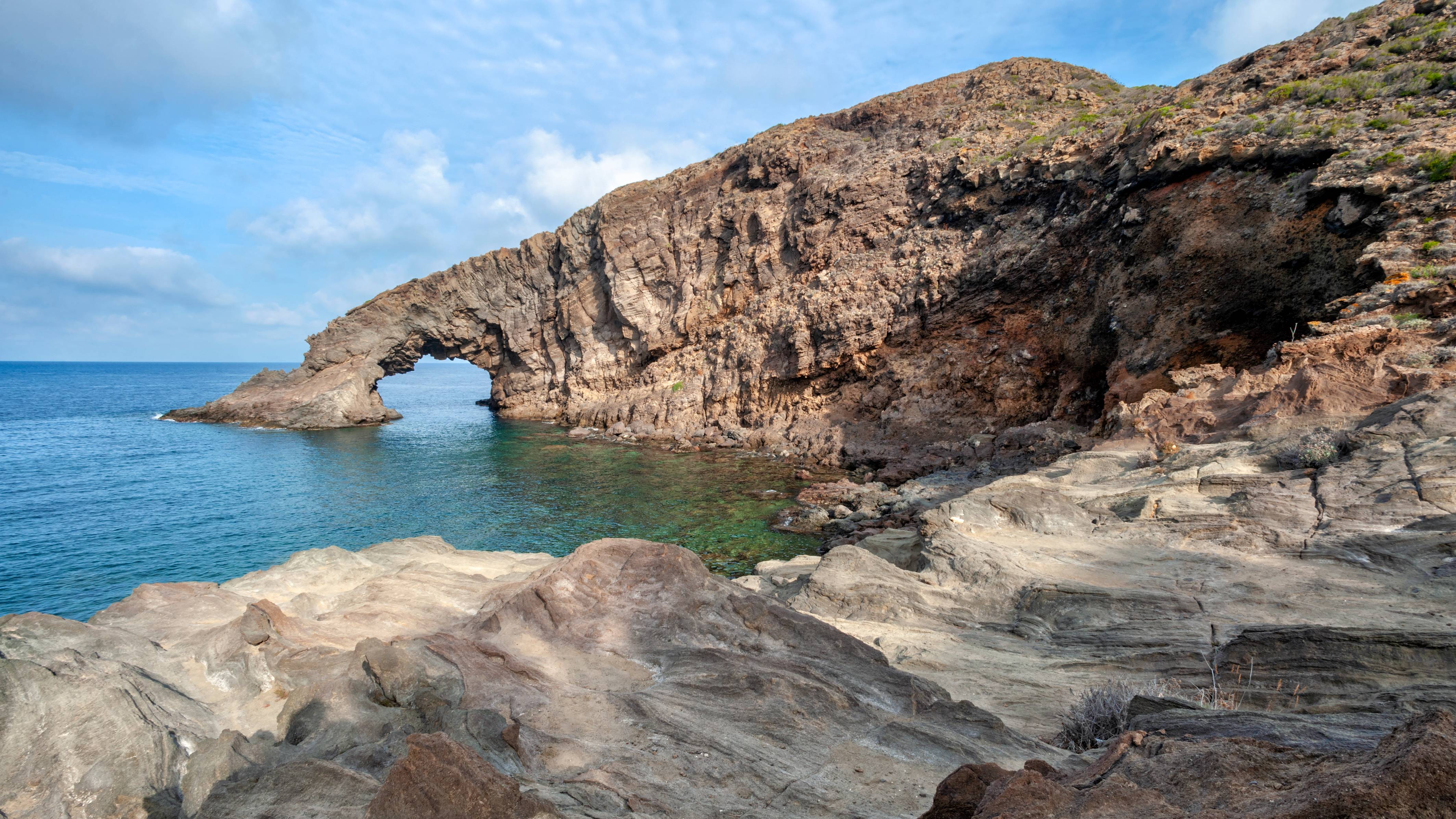 Jagged, rocky foreground leads to a calm, turquoise sea with a large natural arch carved into a cliff face in the background under a partly cloudy sky.