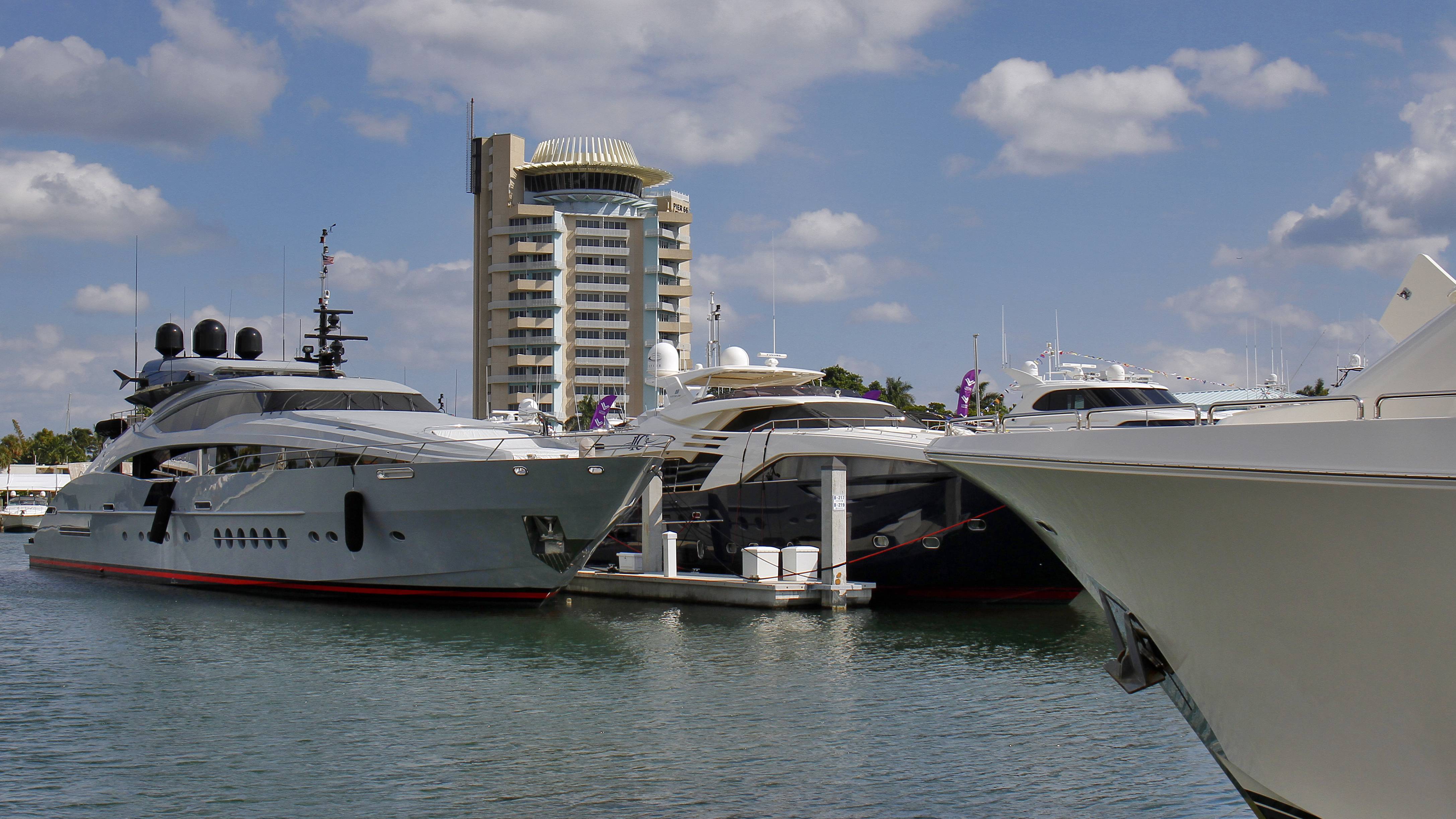 Luxurious yachts are docked in a marina with a distinctive building in the background under a cloudy blue sky.