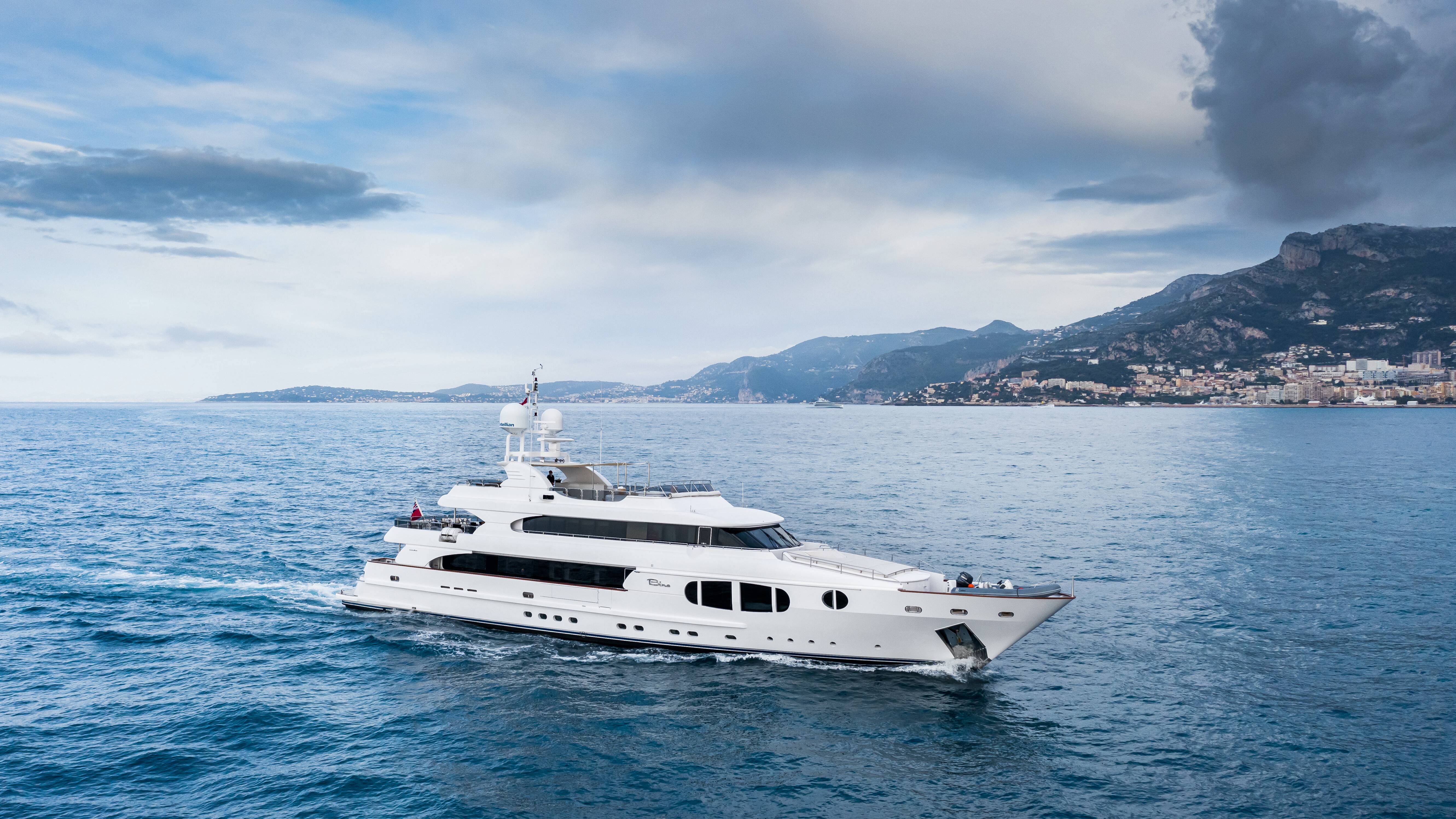 A luxurious white yacht cruises through the deep blue sea with a picturesque coastal town nestled in the mountains behind.