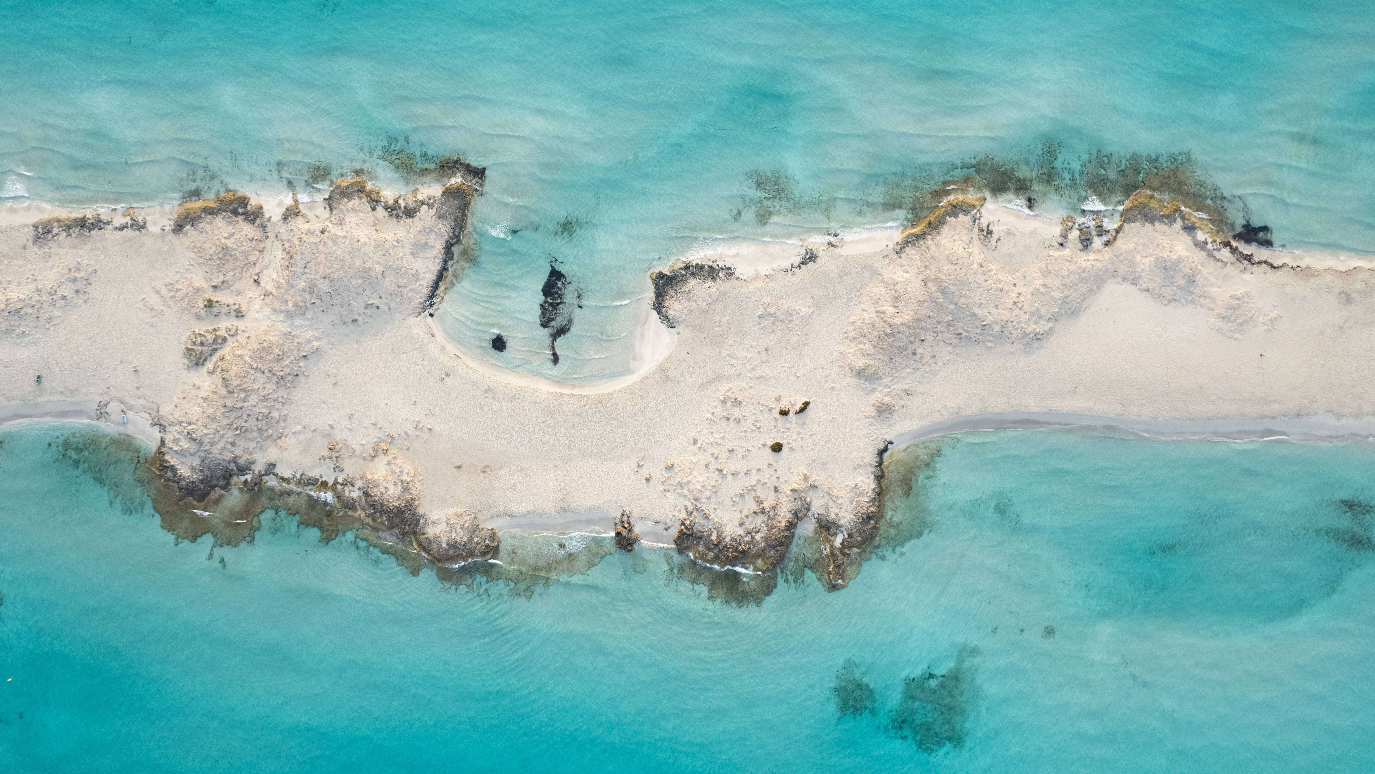 A narrow strip of sandy land with rocky outcrops juts out into the clear turquoise ocean.