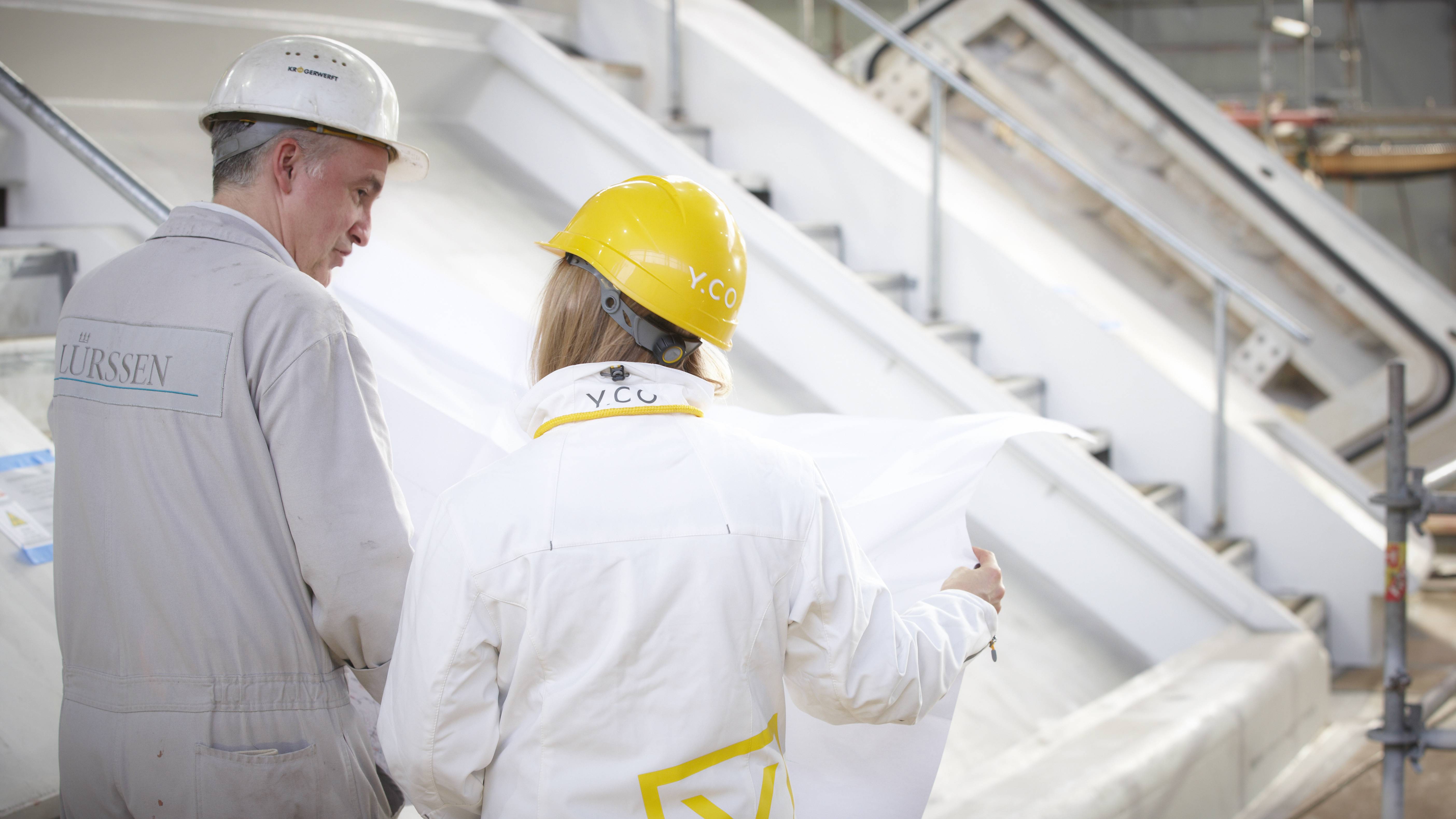Two workers in hard hats review blueprints on a large vessel under construction.