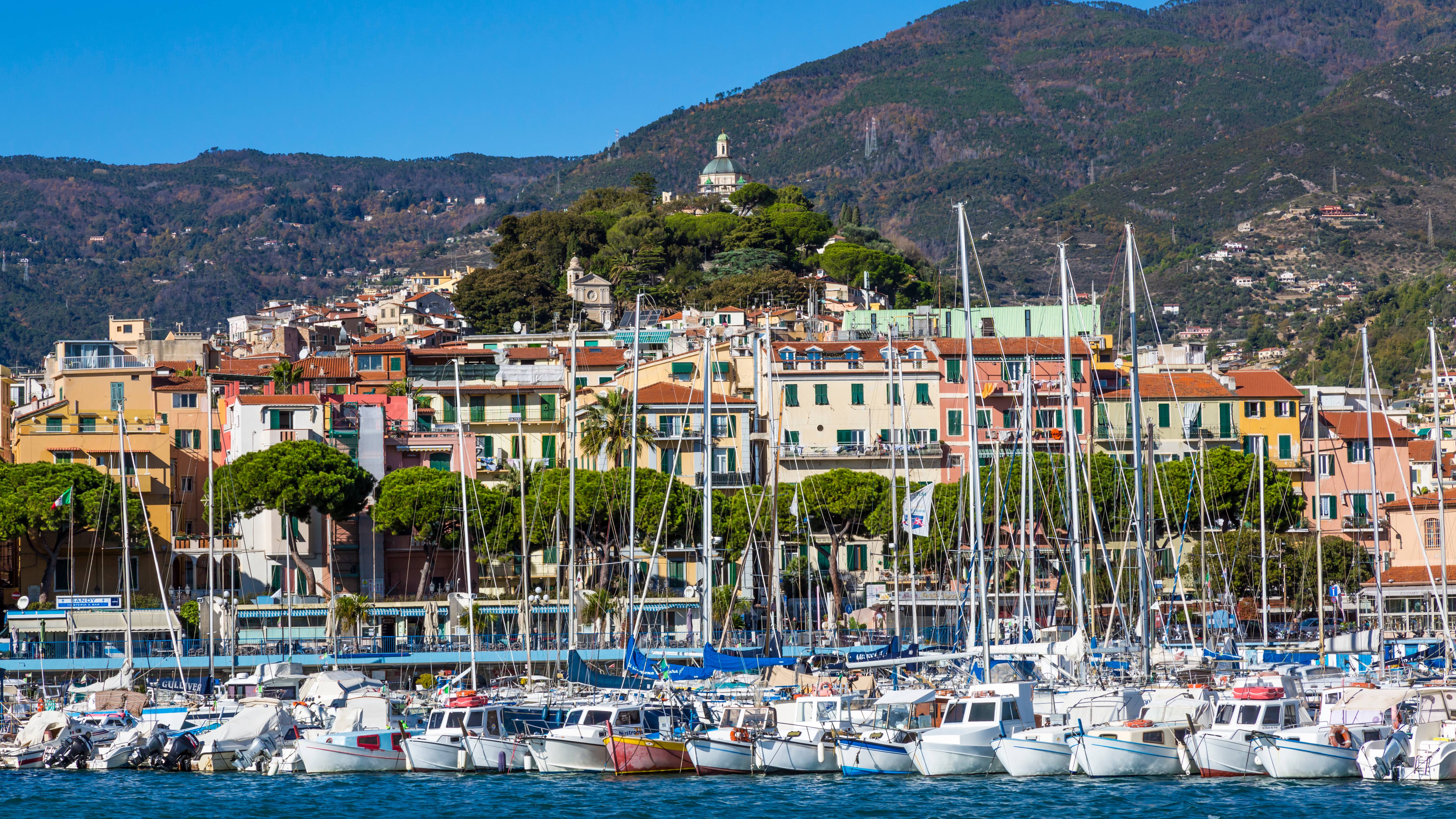 A picturesque Italian harbor bustles with sailboats docked against a backdrop of colorful buildings and a verdant hillside.