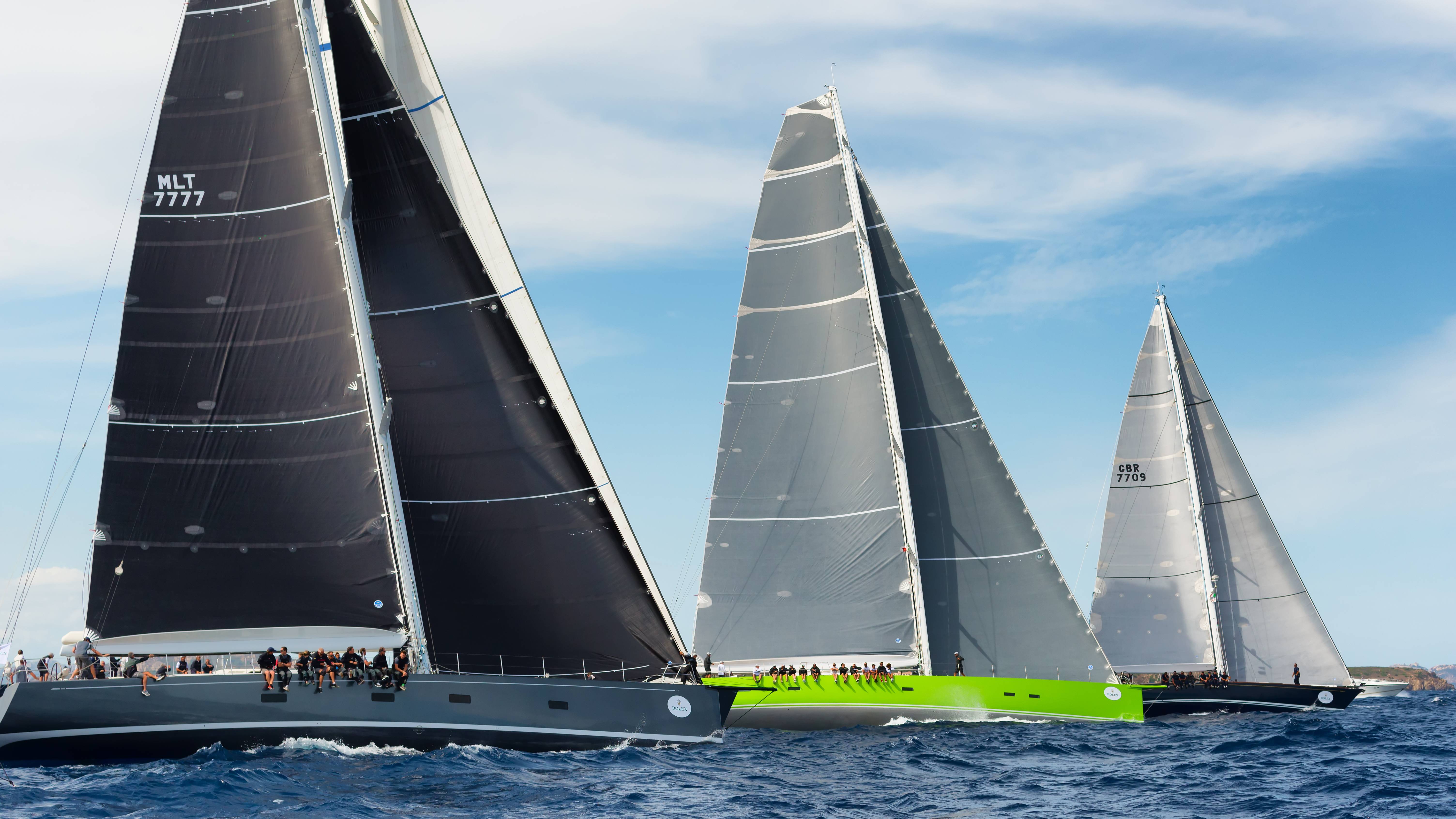 Several large sailboats with dark and light sails race across a blue ocean under a bright sky.