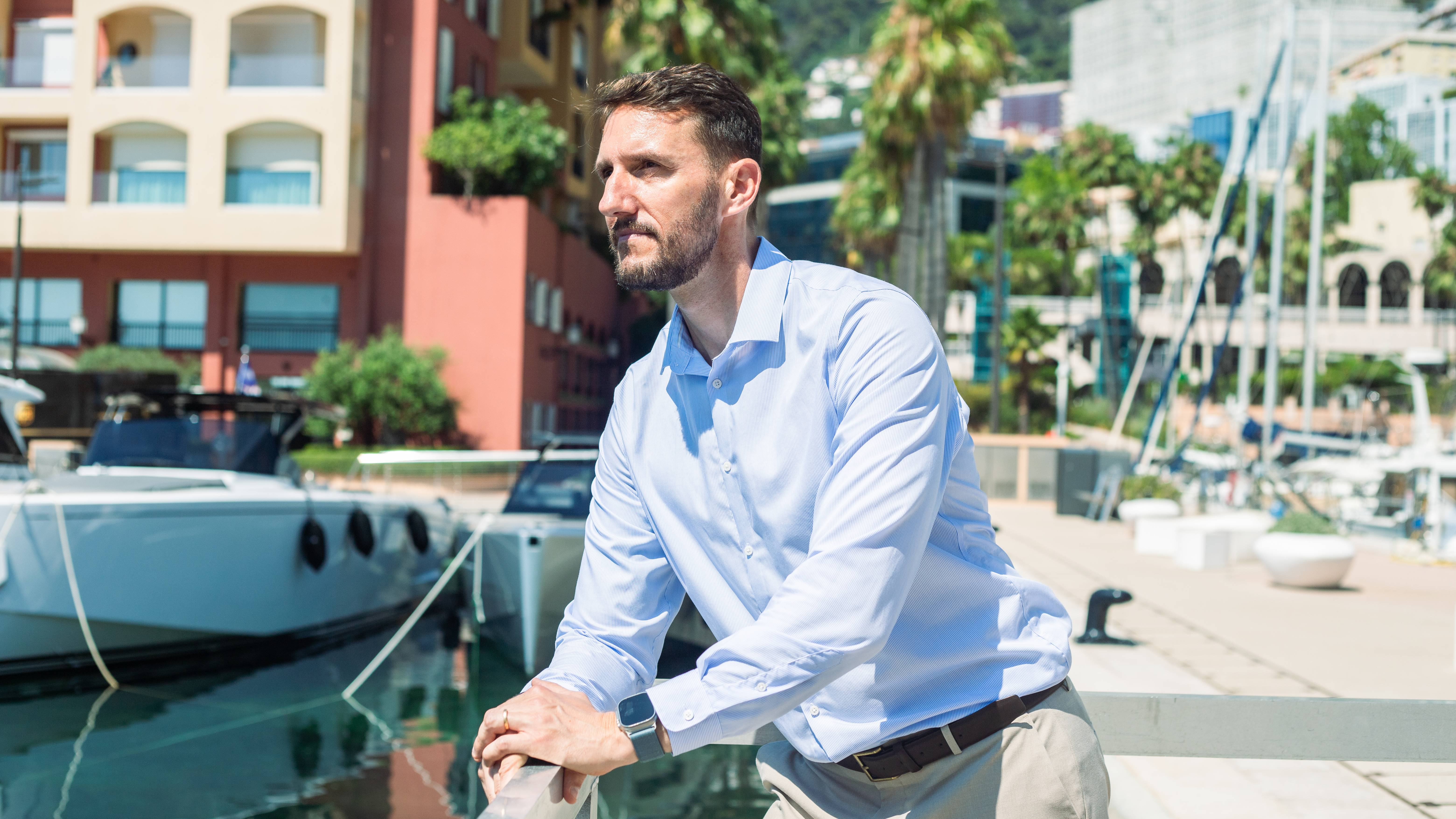 A man in a light blue shirt and tan pants leans on a railing in the foreground, with a harbor filled with boats and buildings in the background.