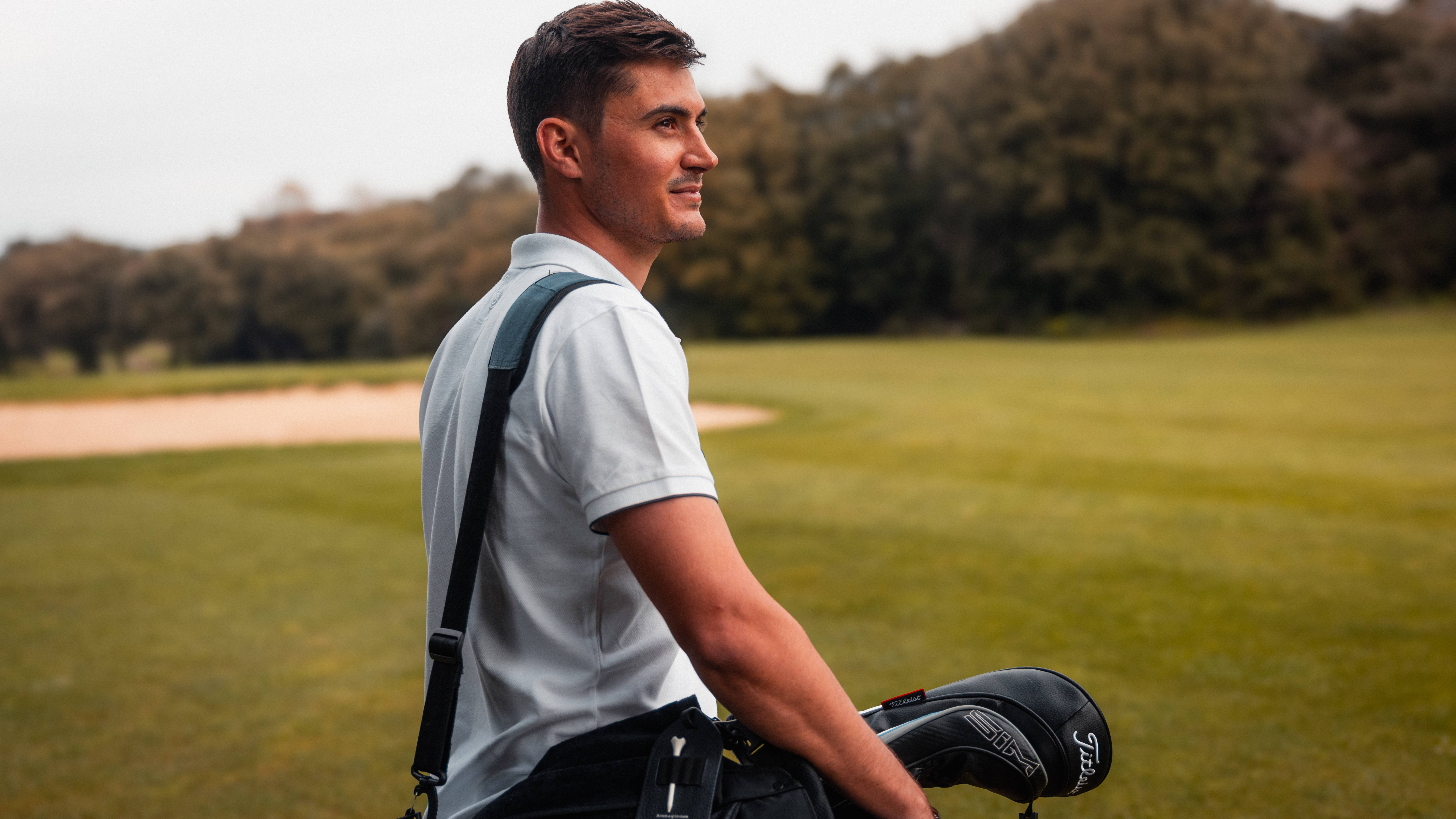 A man in a white polo shirt carries a golf bag across a green course with a sand trap and wooded hills in the background.