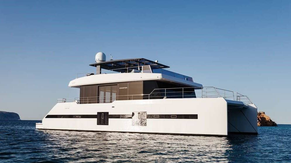 A sleek white catamaran glides through calm blue waters under a clear sky, with rocky islands in the distance.