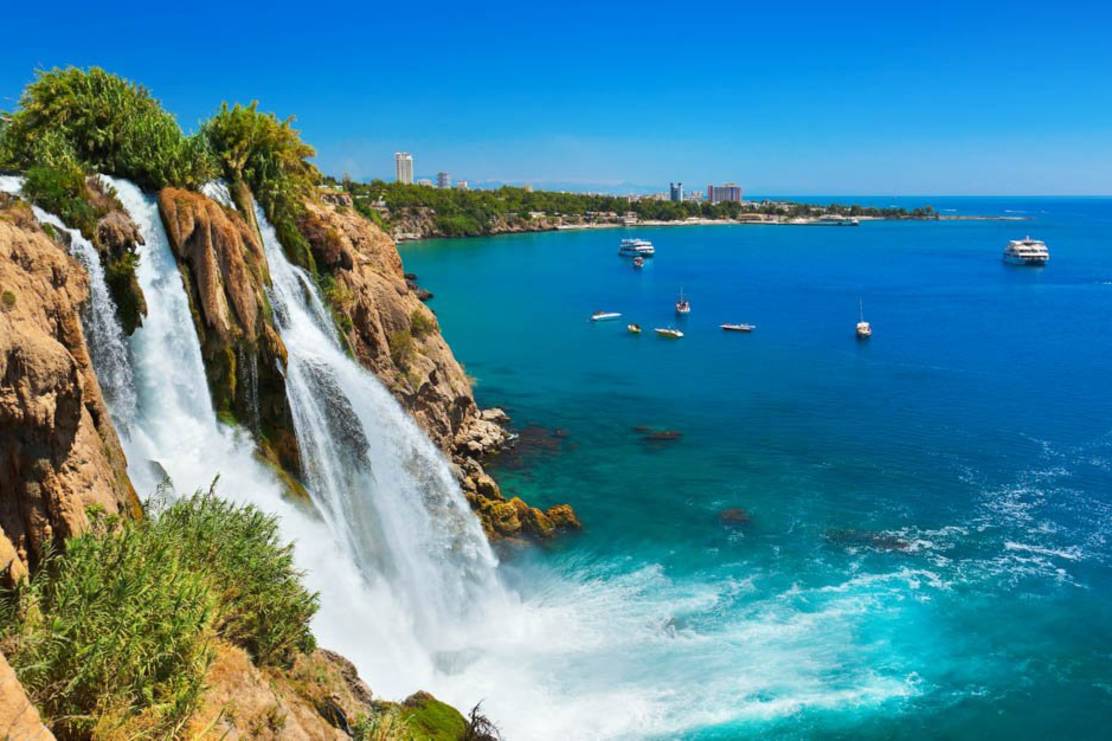 A majestic waterfall cascades down rocky cliffs into the vibrant blue sea, with boats dotting the horizon near a distant city.