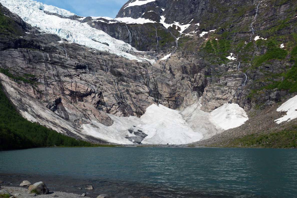 A serene blue lake with rocky shores sits in the foreground, while a massive glacier cascades down a rugged, rocky mountainside in the background.