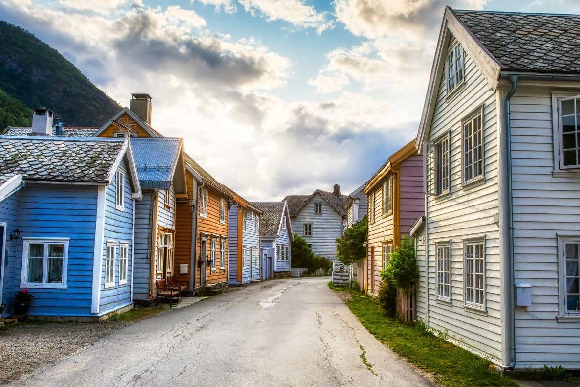 A paved road leads through a village of colorful wooden houses, with a cloudy sky and a mountain visible in the distance.
