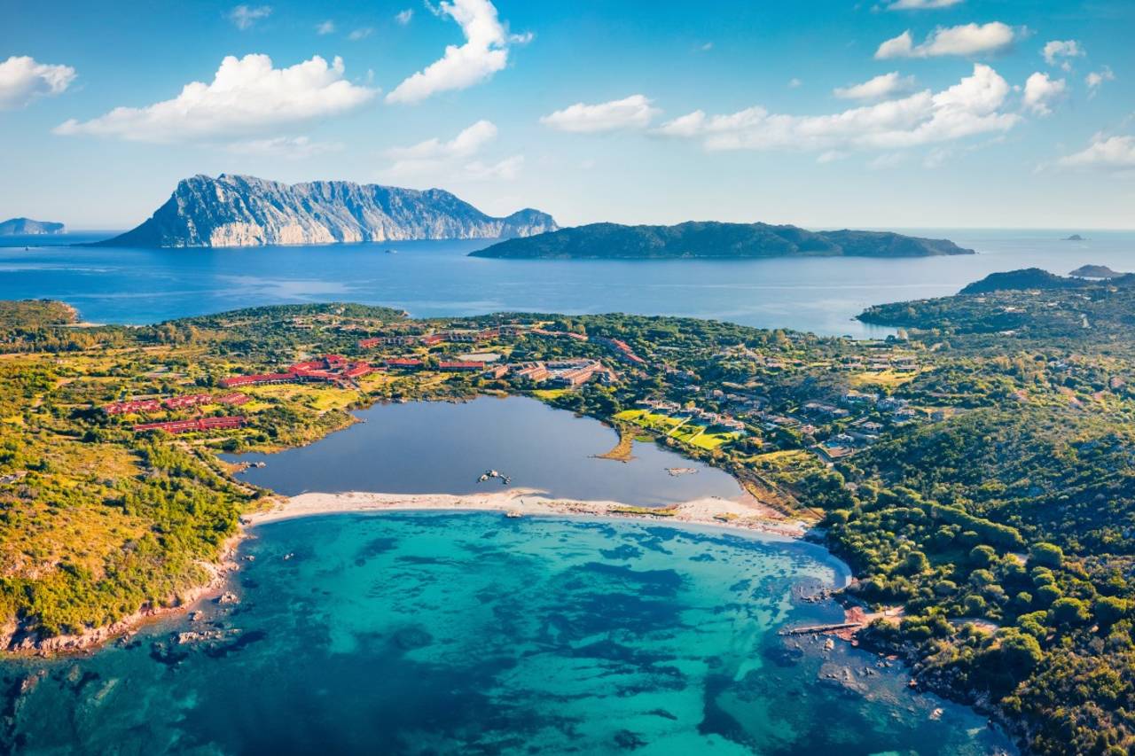 Turquoise waters lap a sandy cove in the foreground, while a serene lake and distant mountainous islands form the background under a bright blue sky.