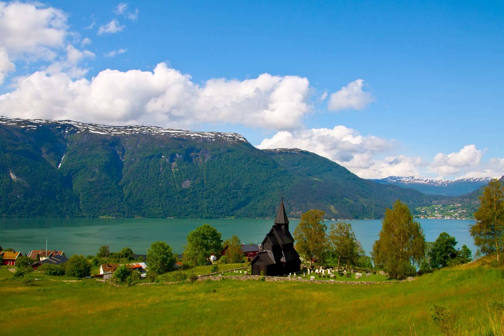 A dark wooden church stands in a grassy field with a lake and mountains in the background.