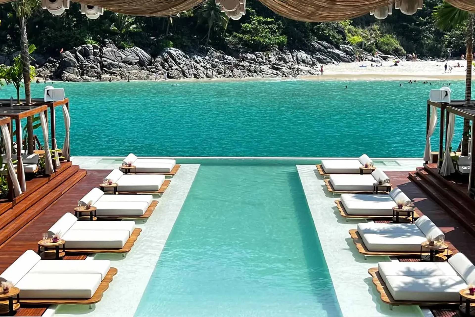 Rows of white lounge chairs surround a bright blue infinity pool in the foreground, with a sandy beach and turquoise ocean stretching into the background.