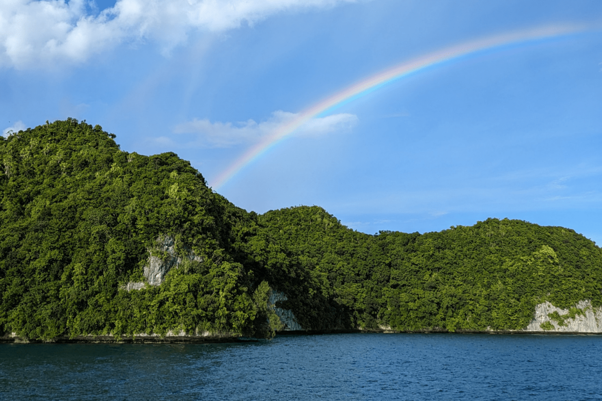 Dark blue water fills the foreground, while lush green islands rise from the sea under a bright blue sky with a rainbow arching across it.