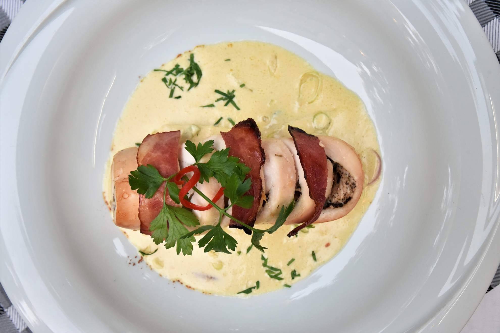 A beautifully plated dish features chicken wrapped in prosciutto, served in a creamy sauce with fresh herbs and a hint of chili.
