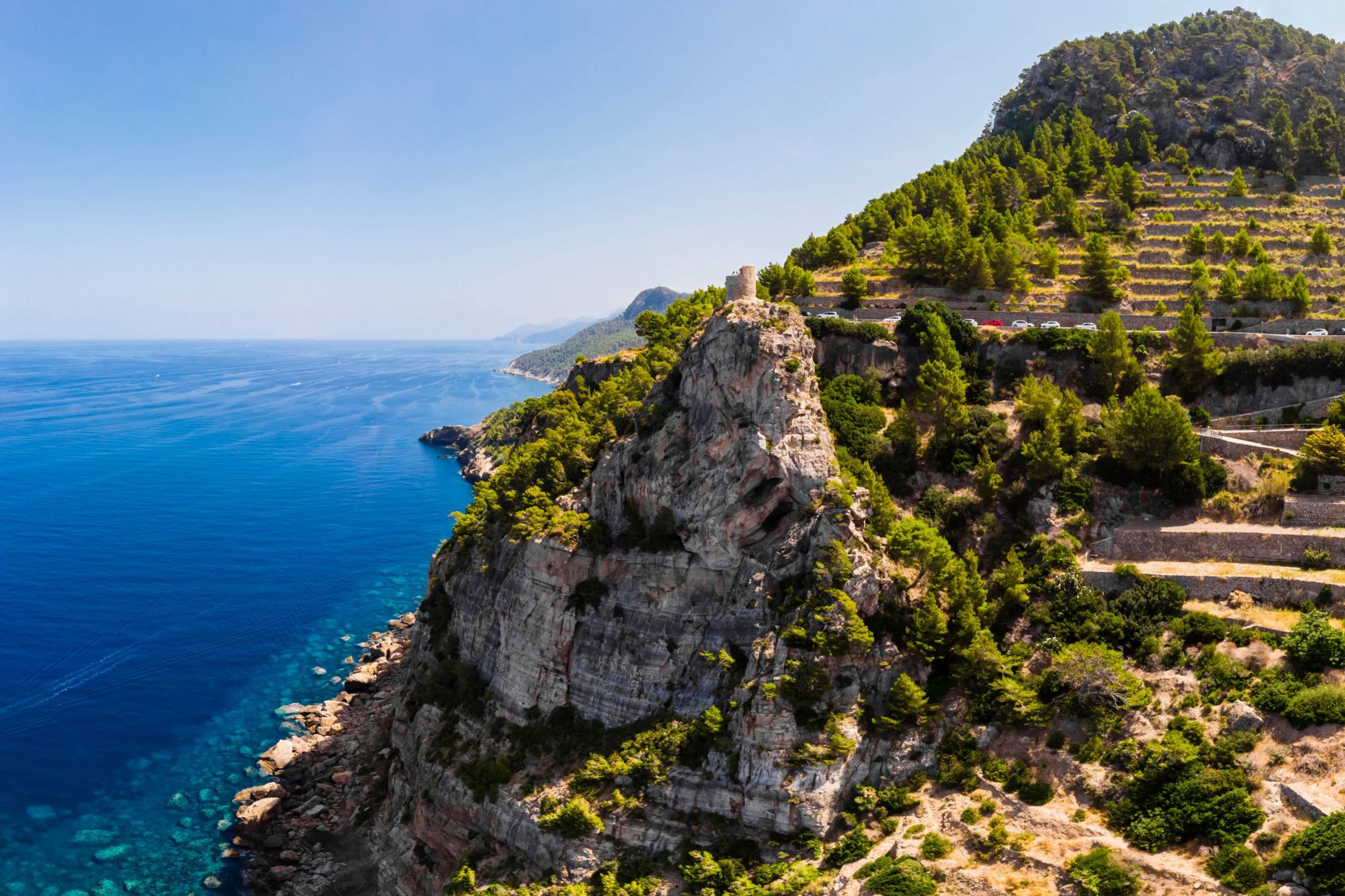 A dramatic cliffside road winds through terraced hillsides overlooking the sparkling blue Mediterranean Sea.