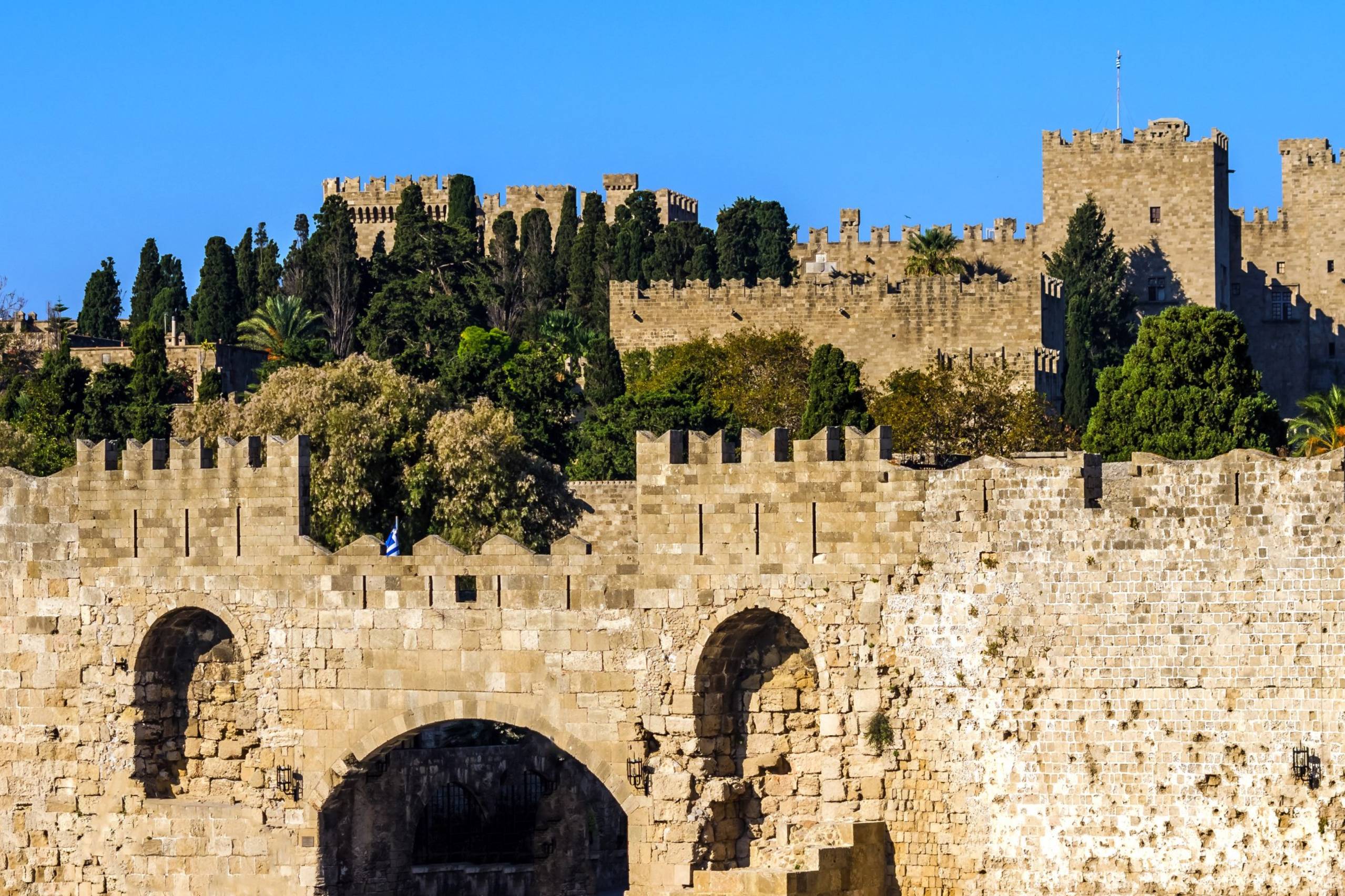 Ancient stone fortifications stand proudly against a clear blue sky, adorned with lush greenery and a distant Greek flag.