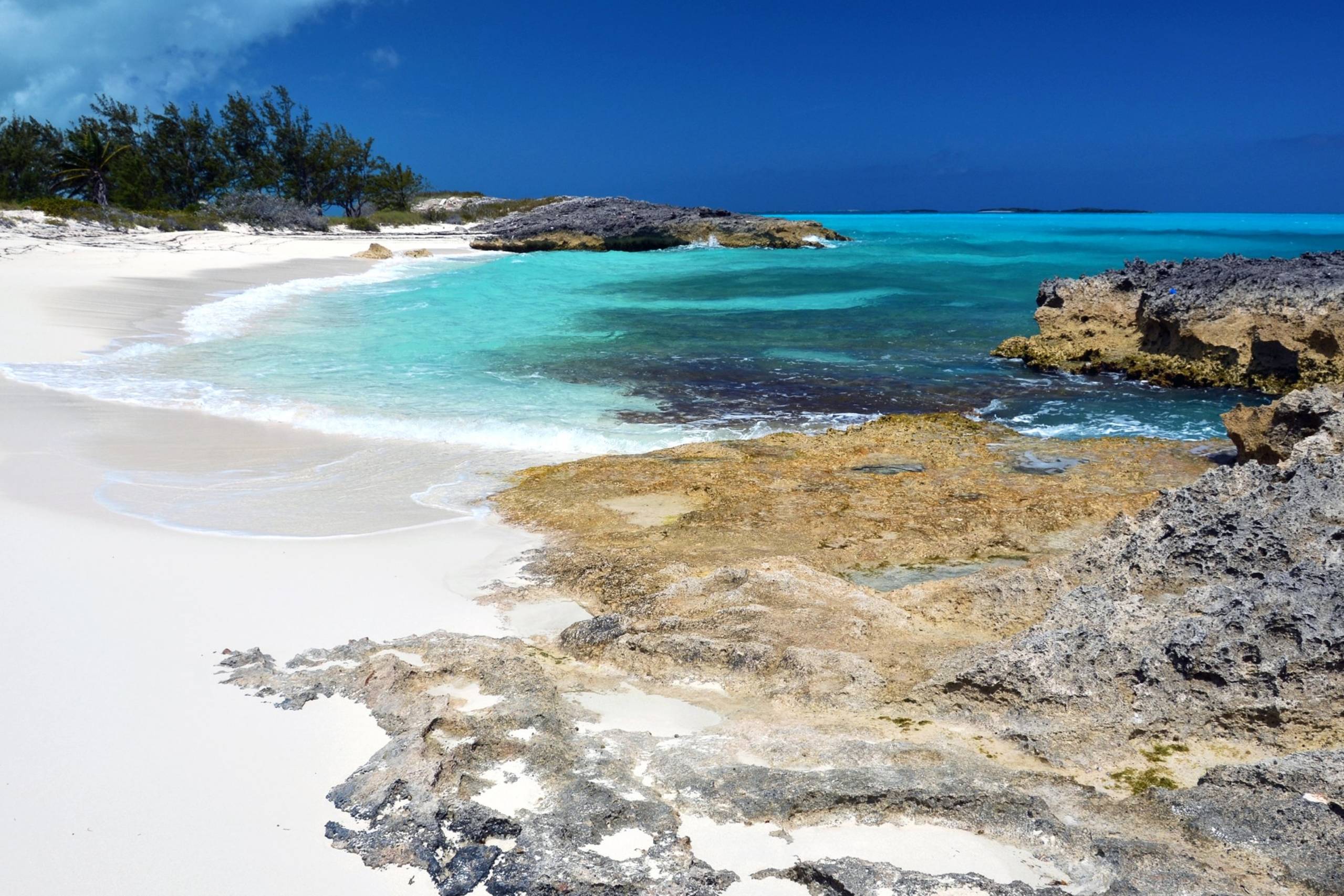 Turquoise waters gently lap onto a pristine white sand beach bordered by rugged coral rock formations under a brilliant blue sky.