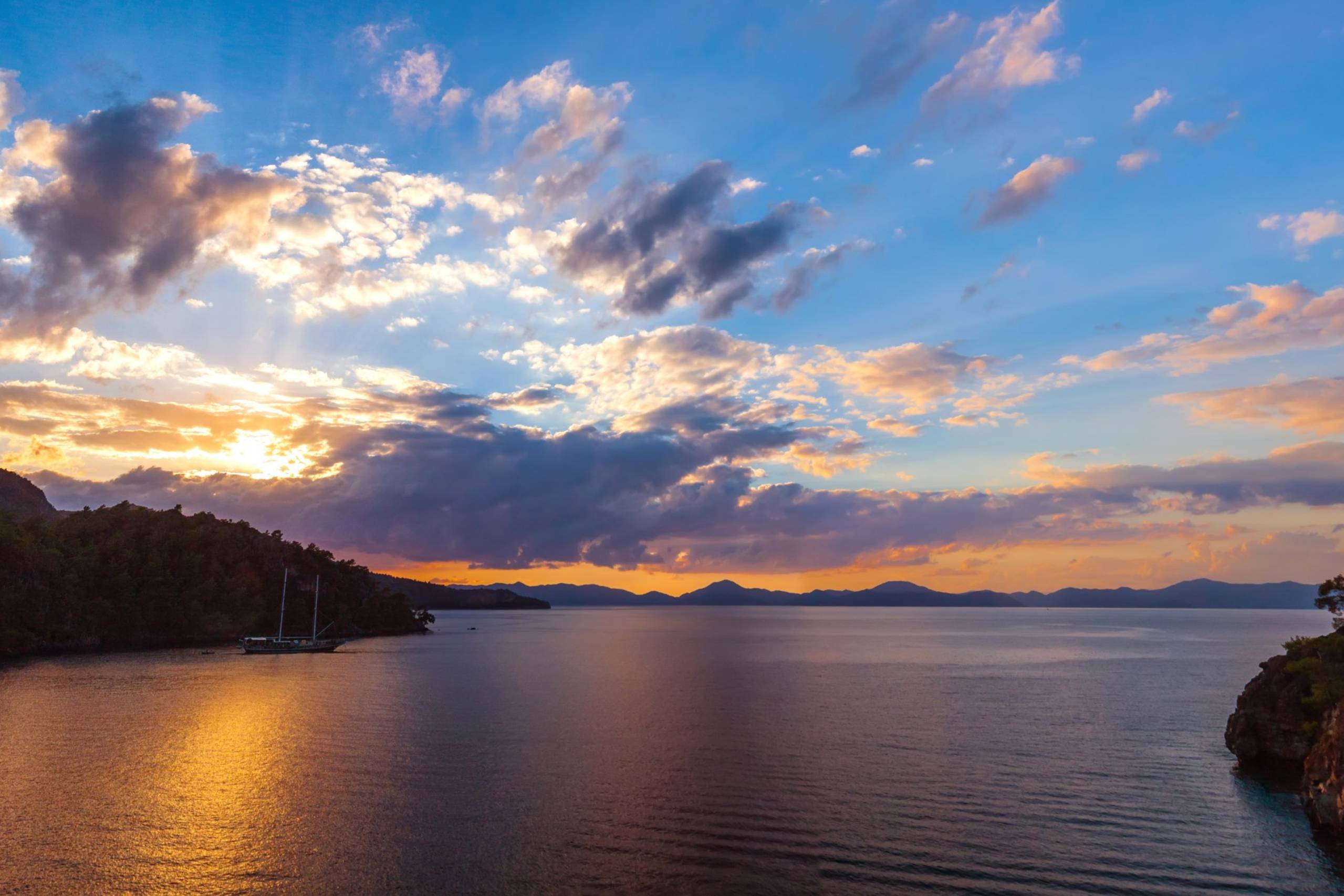 Golden sunset rays illuminate the calm sea and a sailboat anchored near a lush, tree-covered coastline.