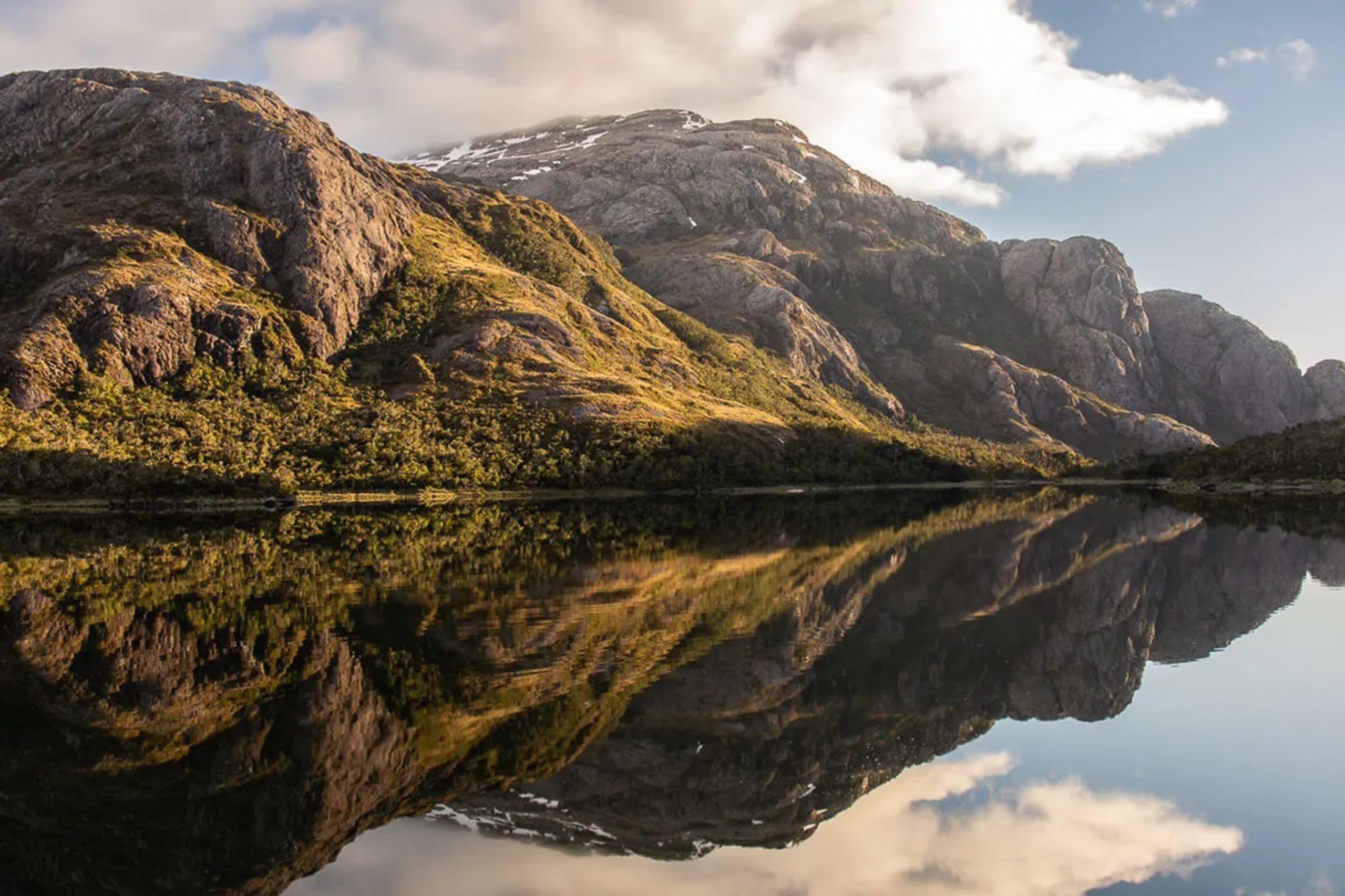 A majestic mountain range is perfectly reflected in the still waters of a serene lake under a cloudy sky.
