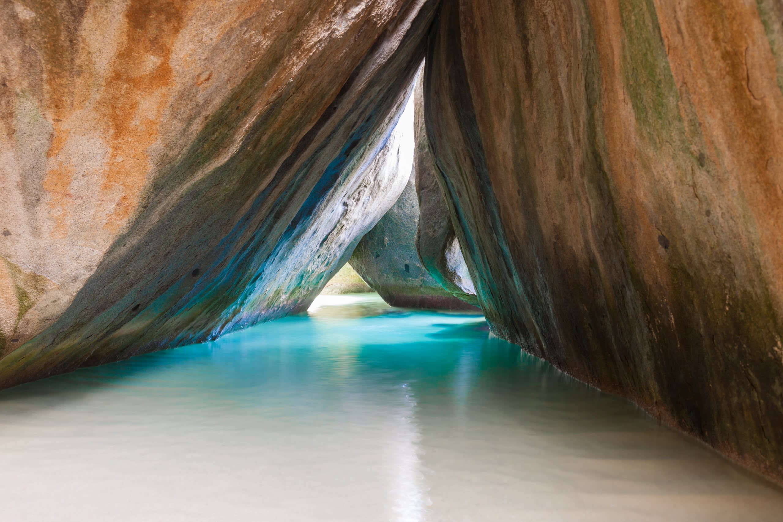 Turquoise waters flow through a natural archway formed by massive, weathered boulders on a tropical shore.