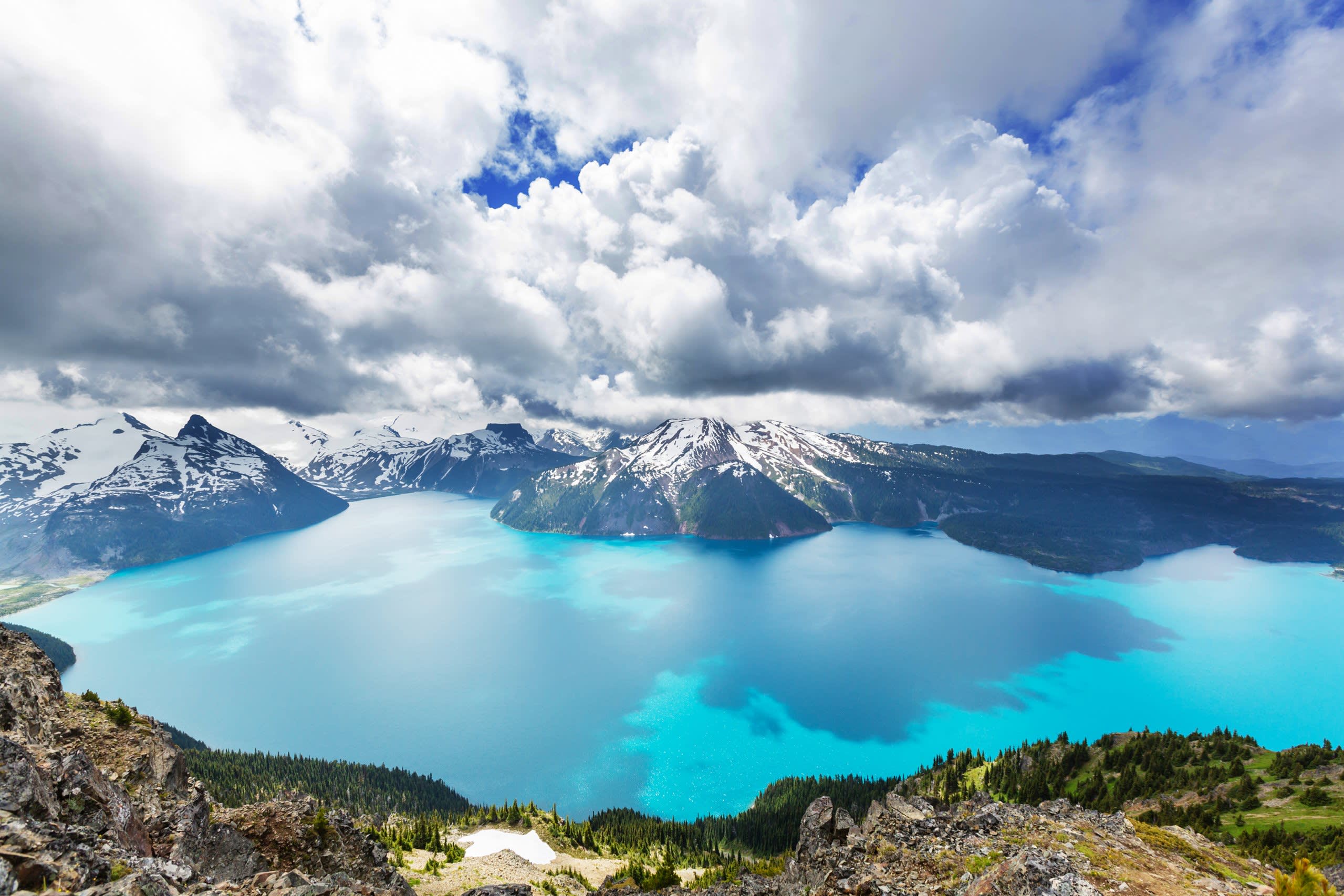 A vast turquoise lake is cradled by snow-capped mountains under a dramatic, cloudy sky.