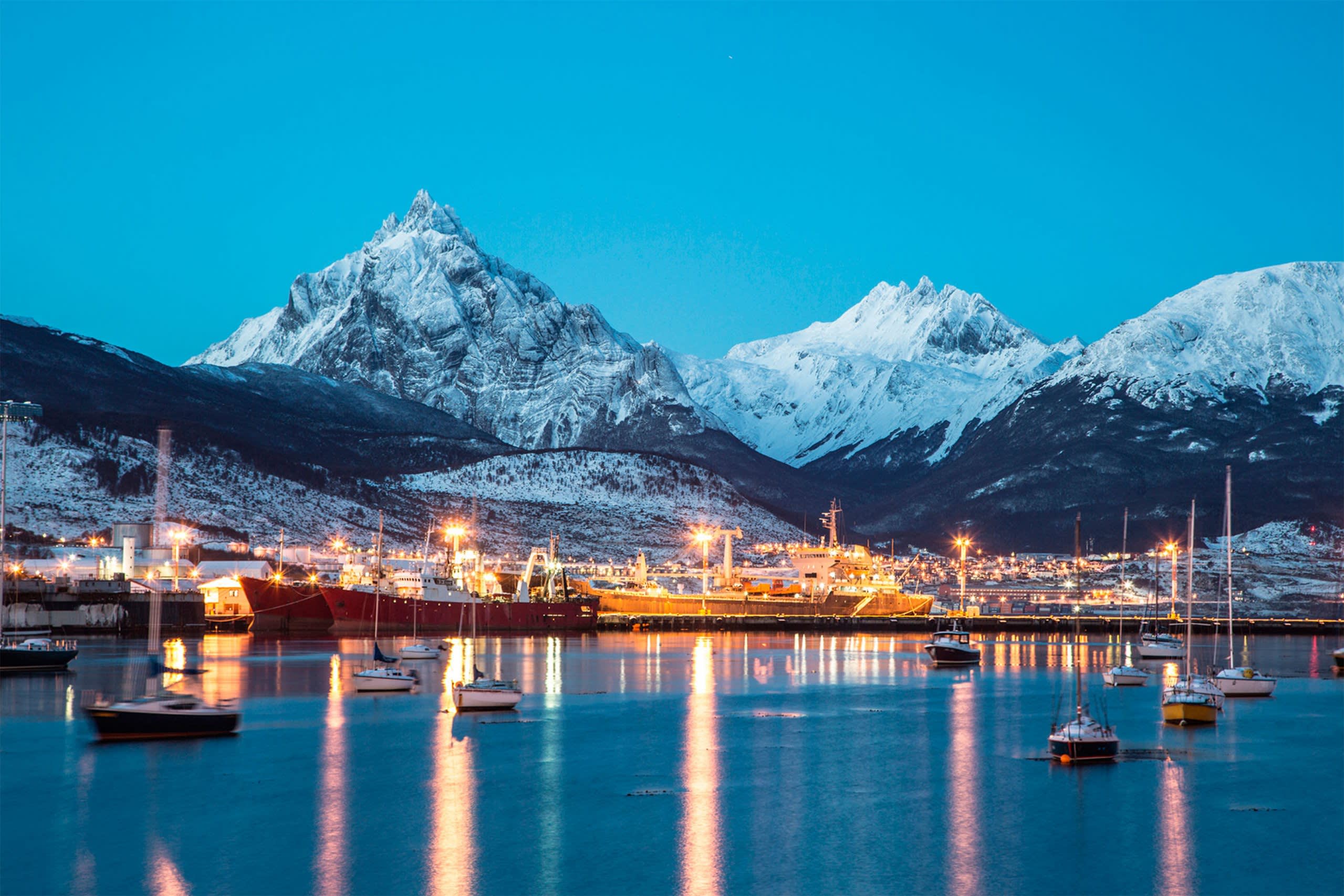 Snow-capped mountains loom over a harbor filled with illuminated ships and sailboats at dusk.