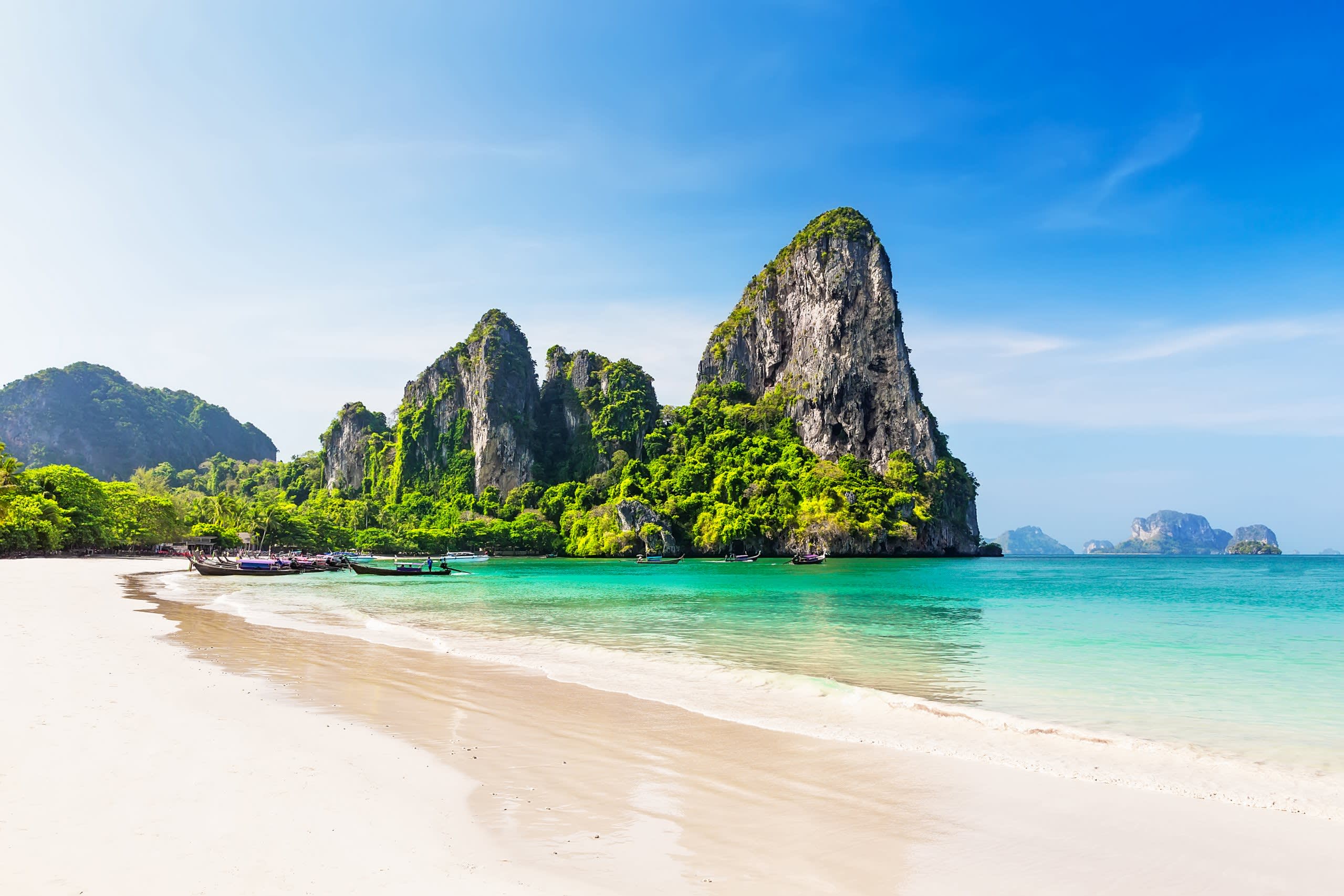 Emerald waters lap a pristine white sand beach at the base of lush, jungle-covered limestone karsts under a brilliant blue sky.
