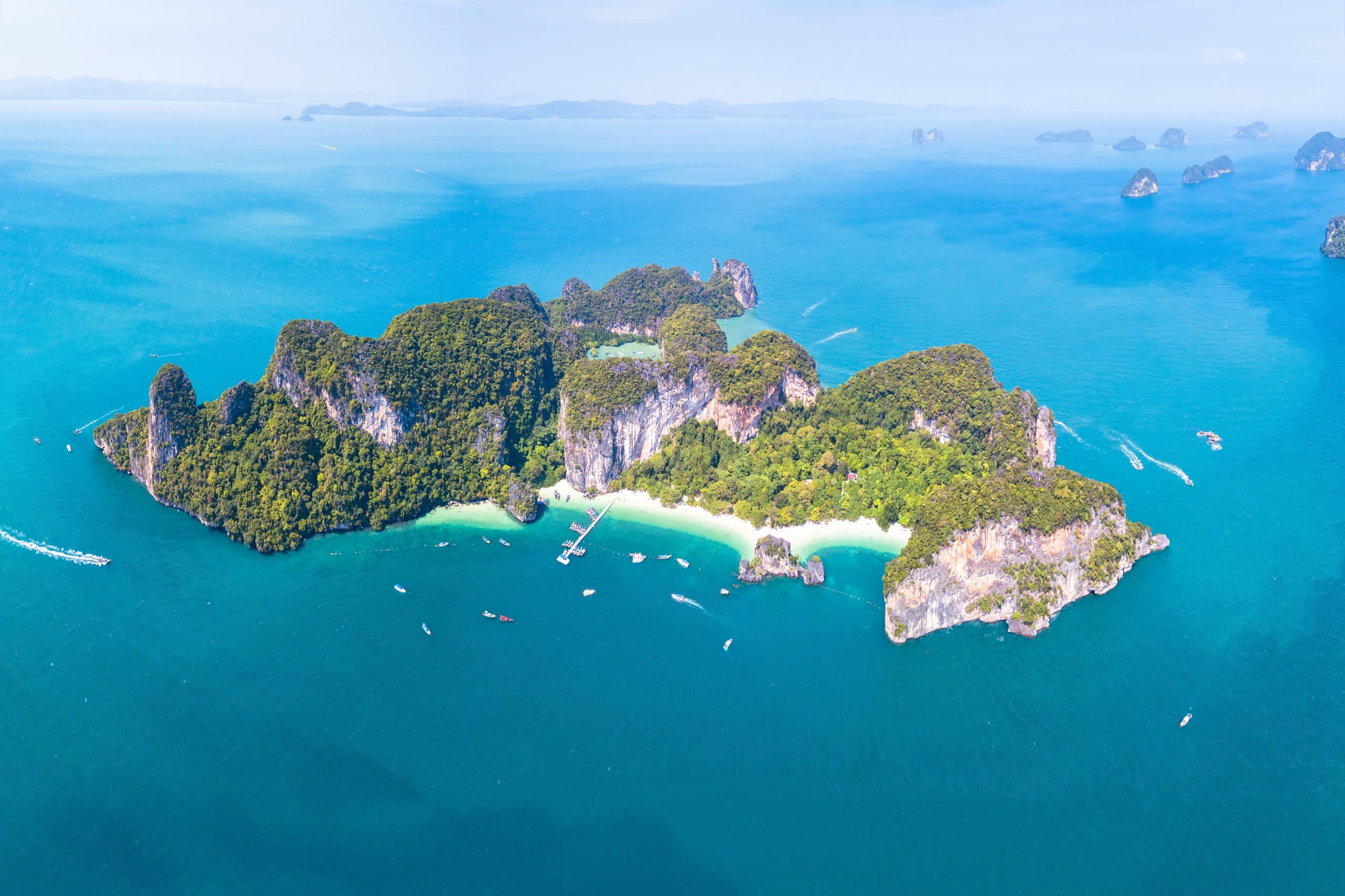Lush green islands rise from the turquoise waters of Thailand, dotted with boats and pristine white-sand beaches.