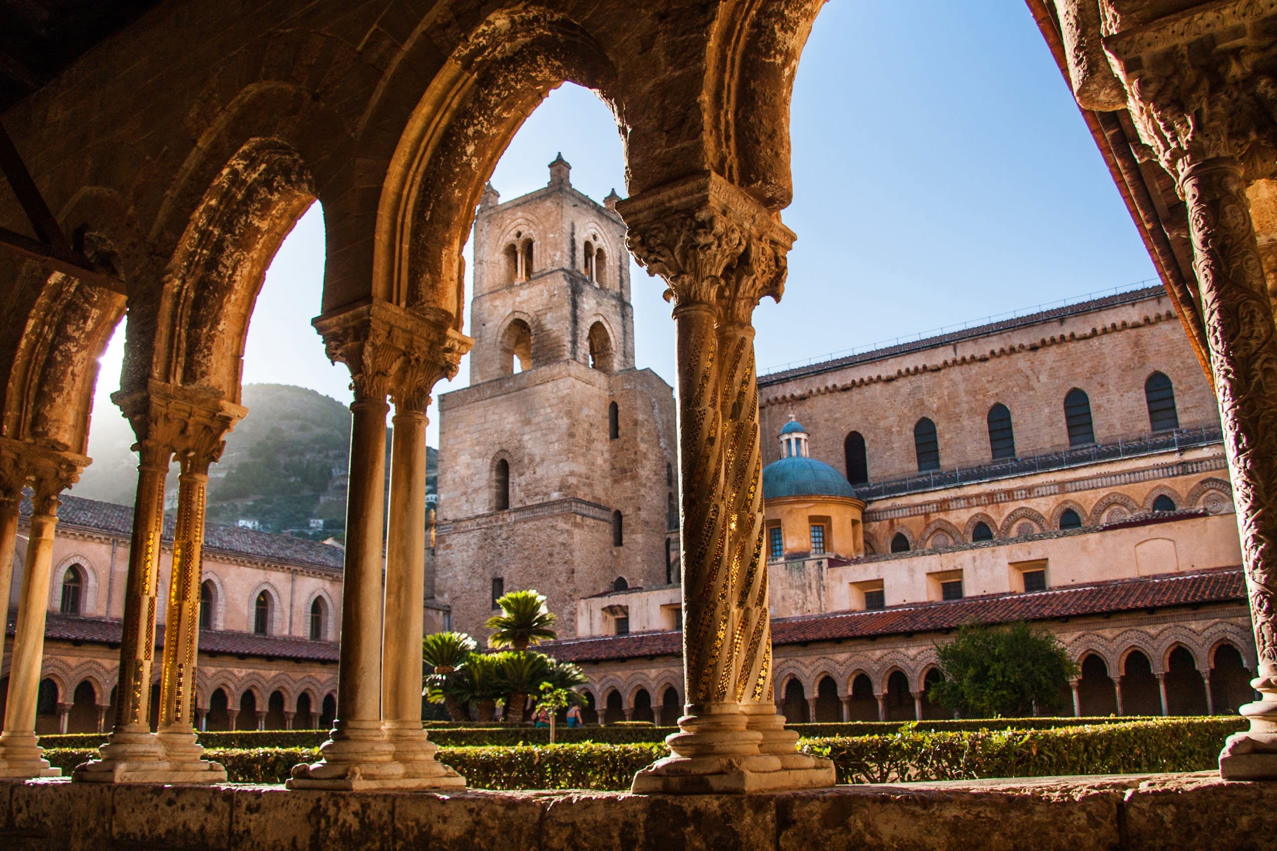 Sunlight illuminates the ornate cloister of a historic cathedral, framing a view of its bell tower and surrounding architecture.