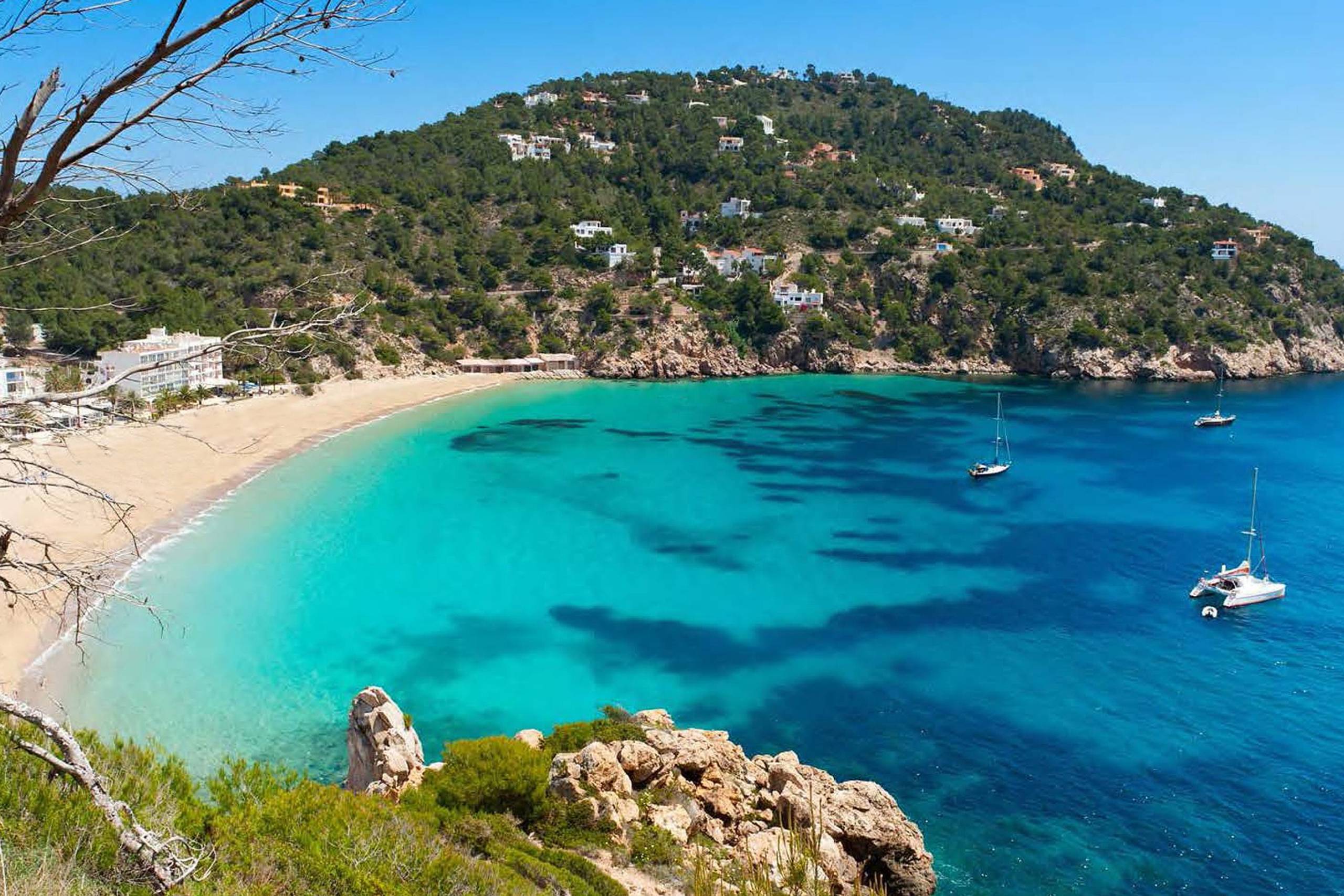 A secluded cove with turquoise waters, a sandy beach, and villas nestled into the lush green hillside offers a serene Mediterranean escape.