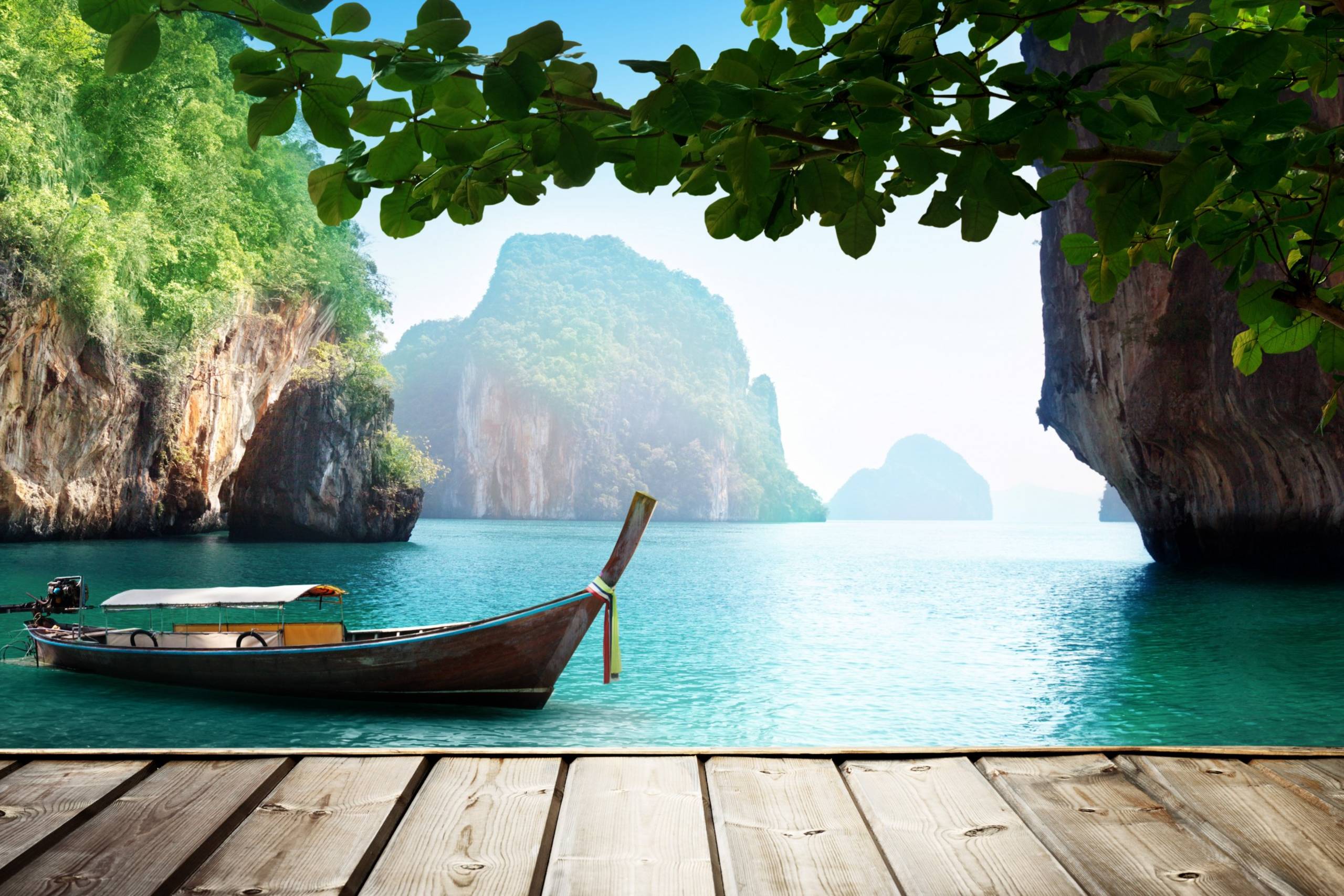 A traditional longtail boat floats serenely in the turquoise waters of a tropical bay, framed by lush green foliage and dramatic limestone cliffs.