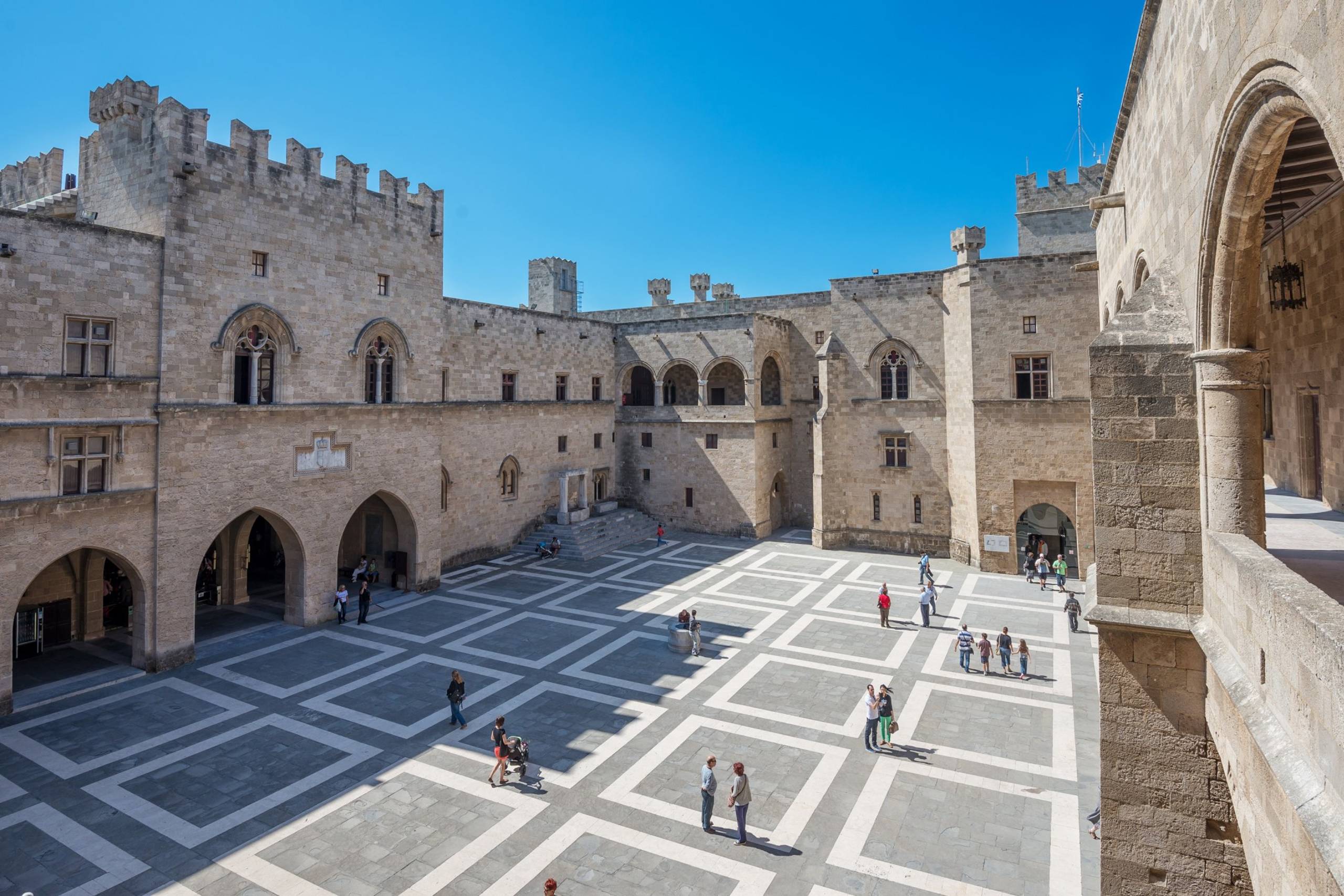Tourists explore the vast courtyard of the Palace of the Grand Master of the Knights of Rhodes under a clear blue sky.