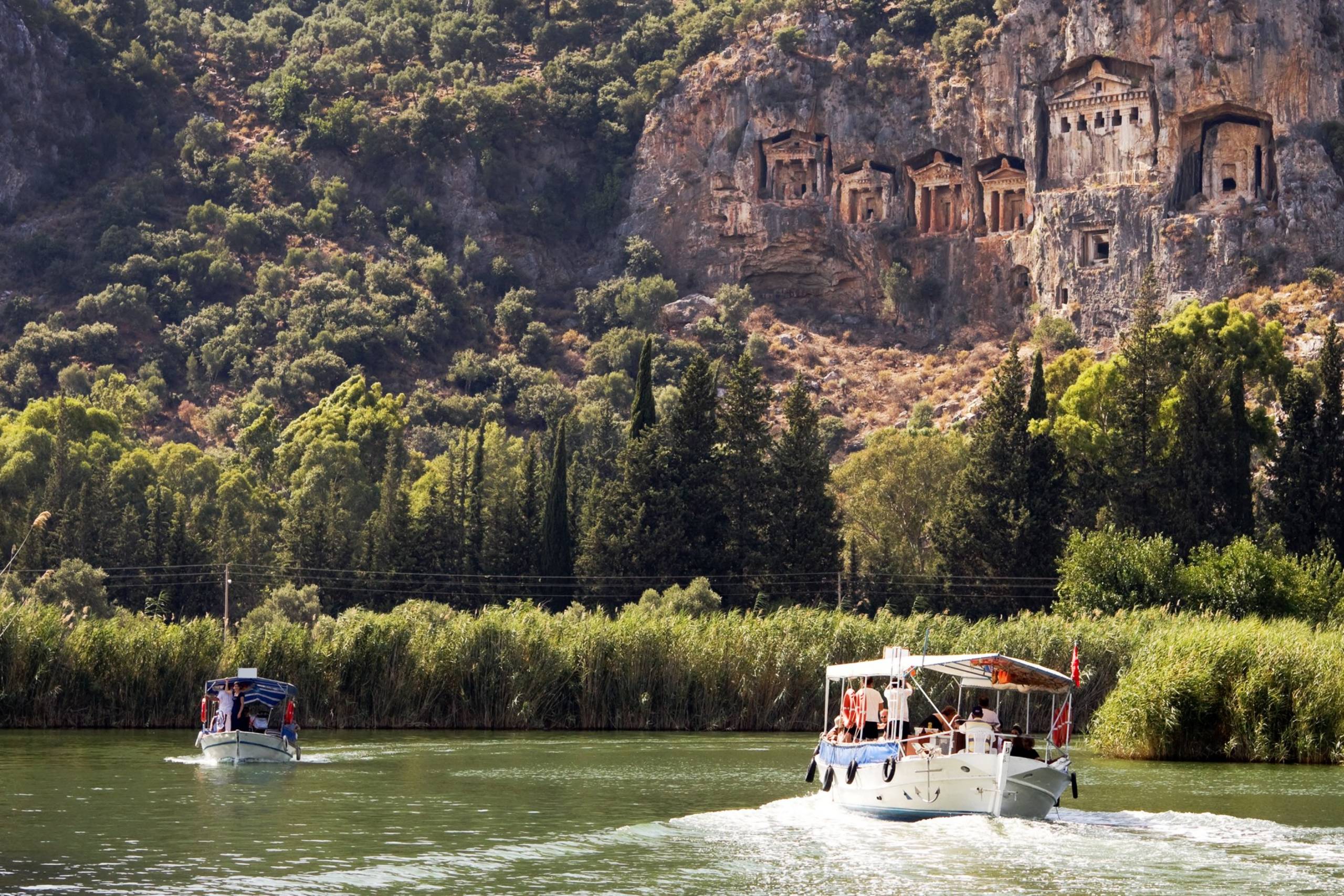 Tourists on a boat cruise past ancient Lycian rock tombs carved into a towering cliff face.