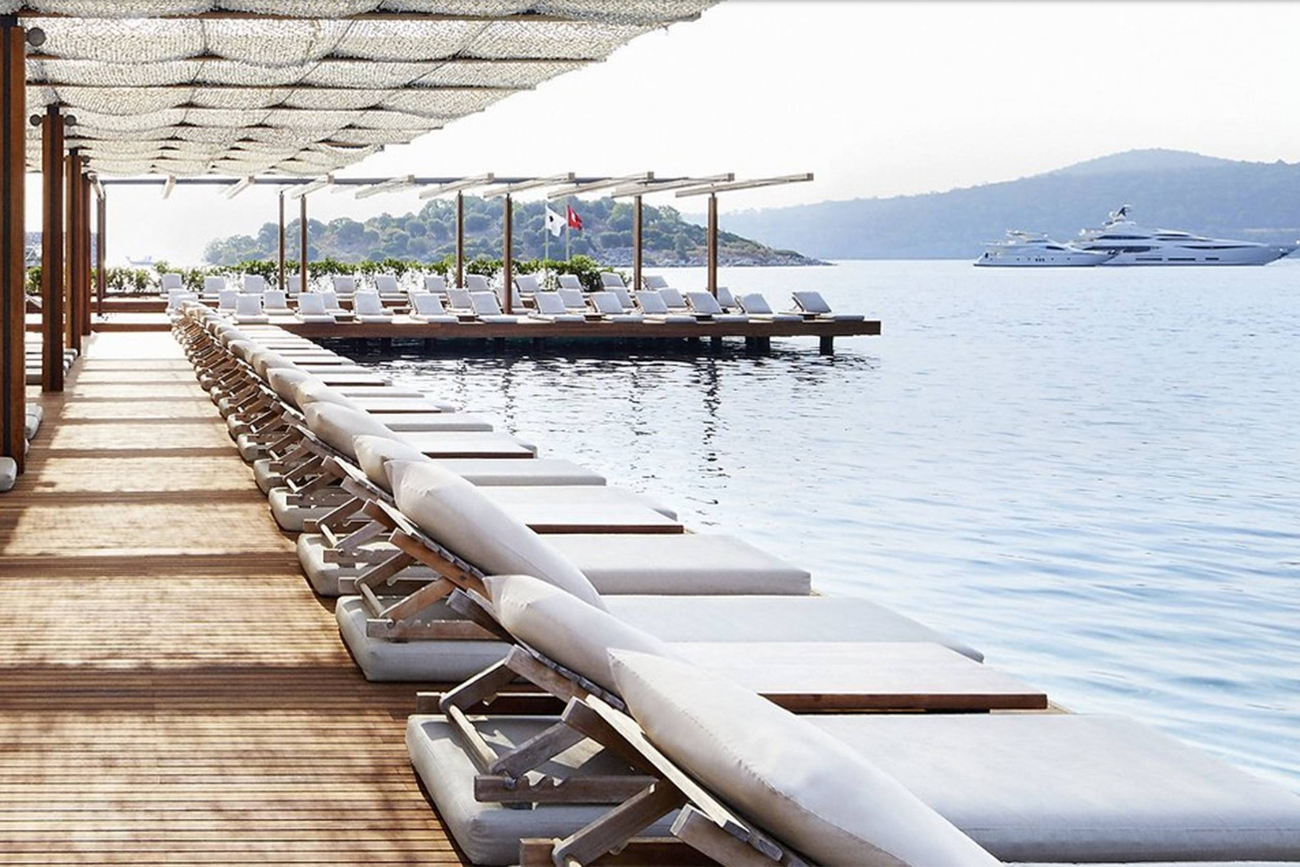 Luxurious lounge chairs line a wooden deck overlooking the tranquil blue water and a distant yacht.