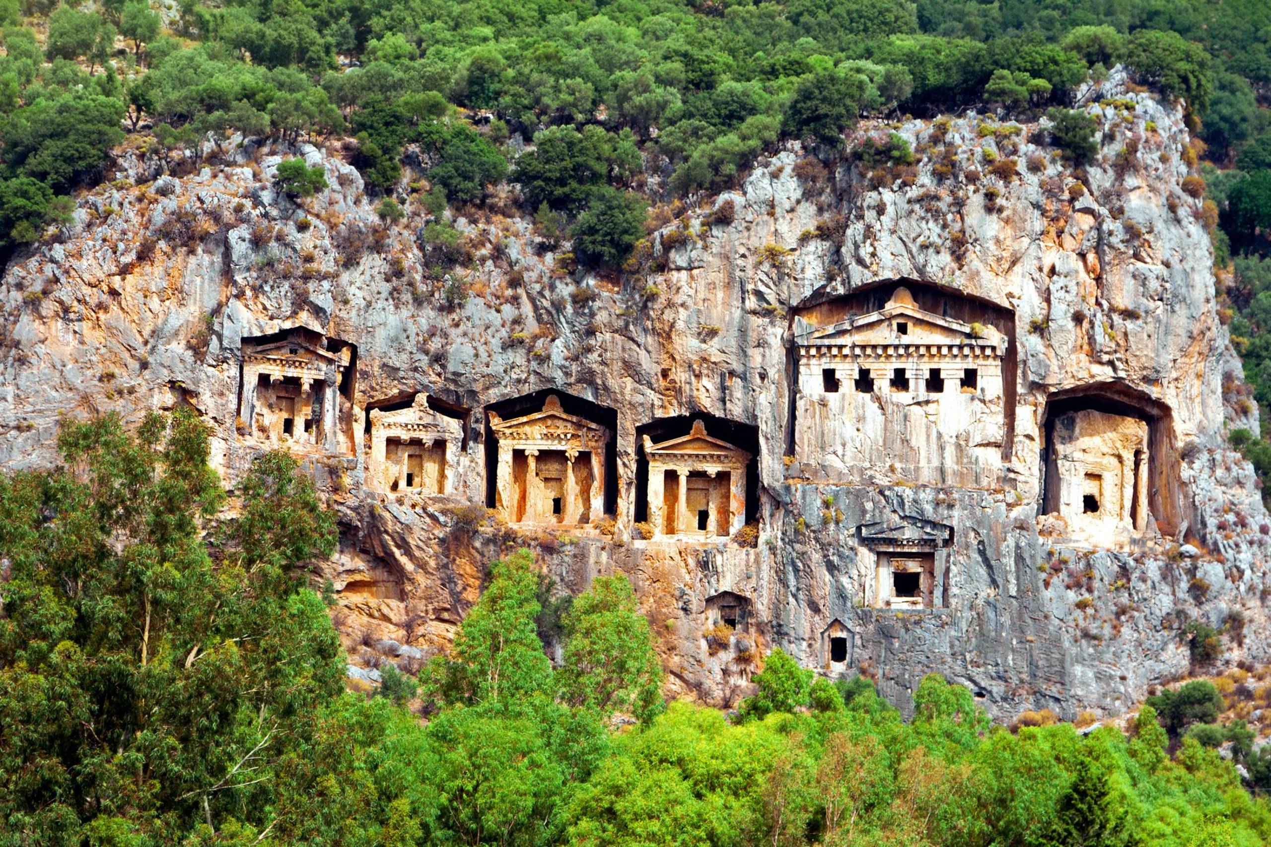 Ancient Lycian rock tombs carved into a sheer cliff face are nestled amongst lush green trees.