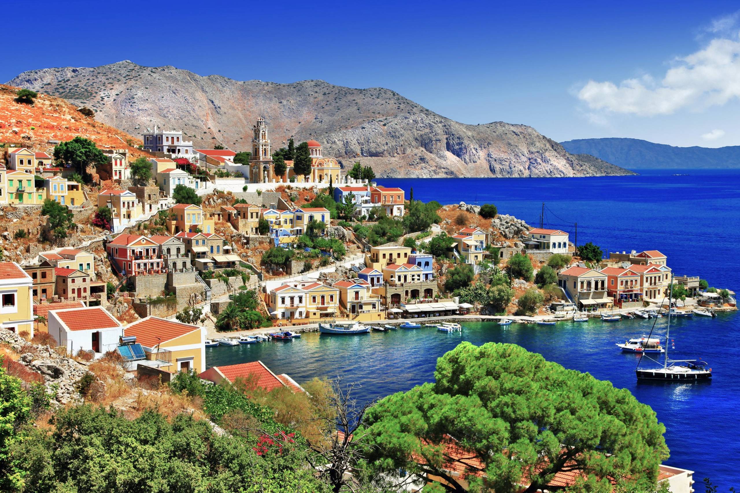 Colorful houses cascade down a hillside to a vibrant blue bay dotted with sailboats.