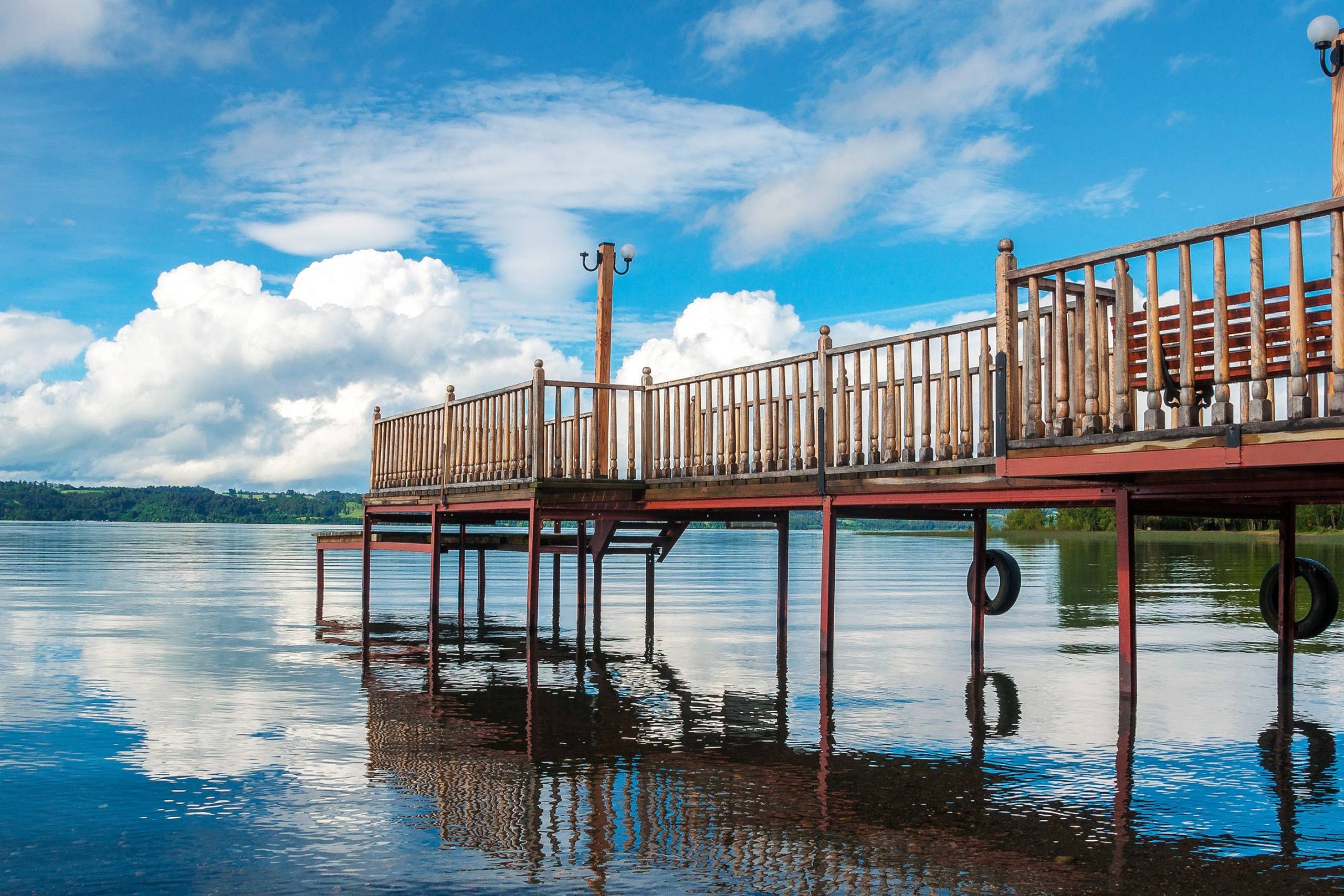 A wooden pier extends over a tranquil lake, reflecting the vibrant blue sky and fluffy white clouds.