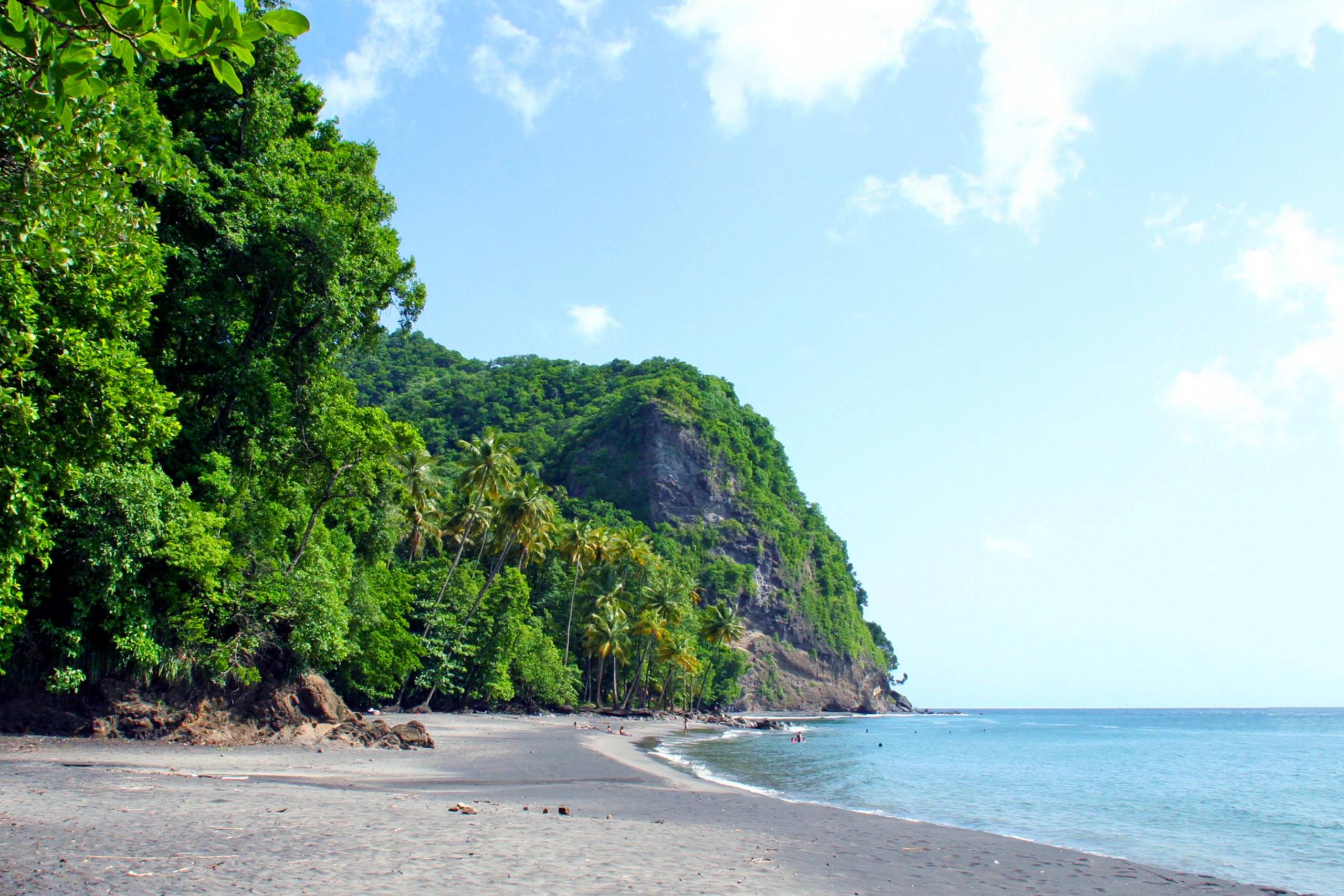 Lush green cliffs meet a dark sand beach and tranquil turquoise waters under a bright blue sky.