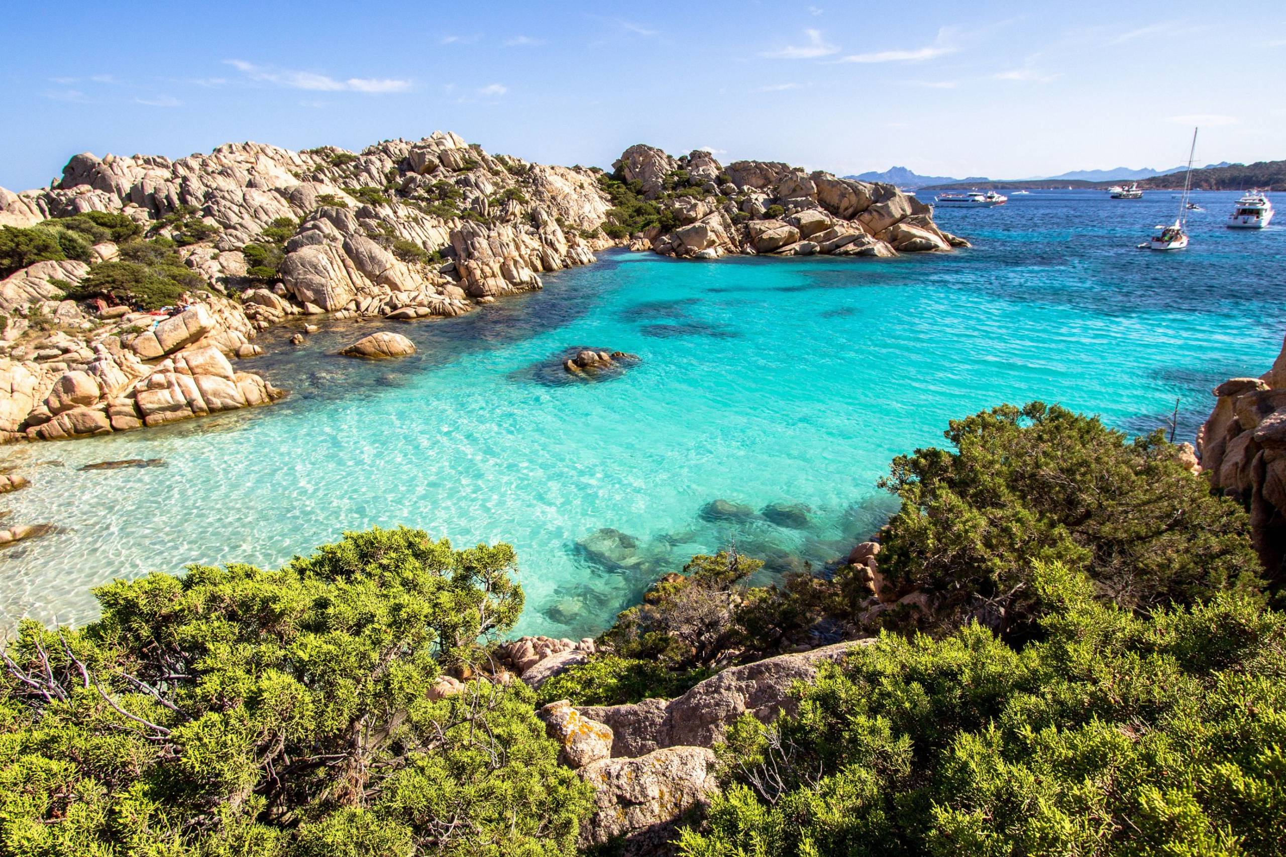 Turquoise waters lap against a rocky cove dotted with green shrubs under a clear blue sky.