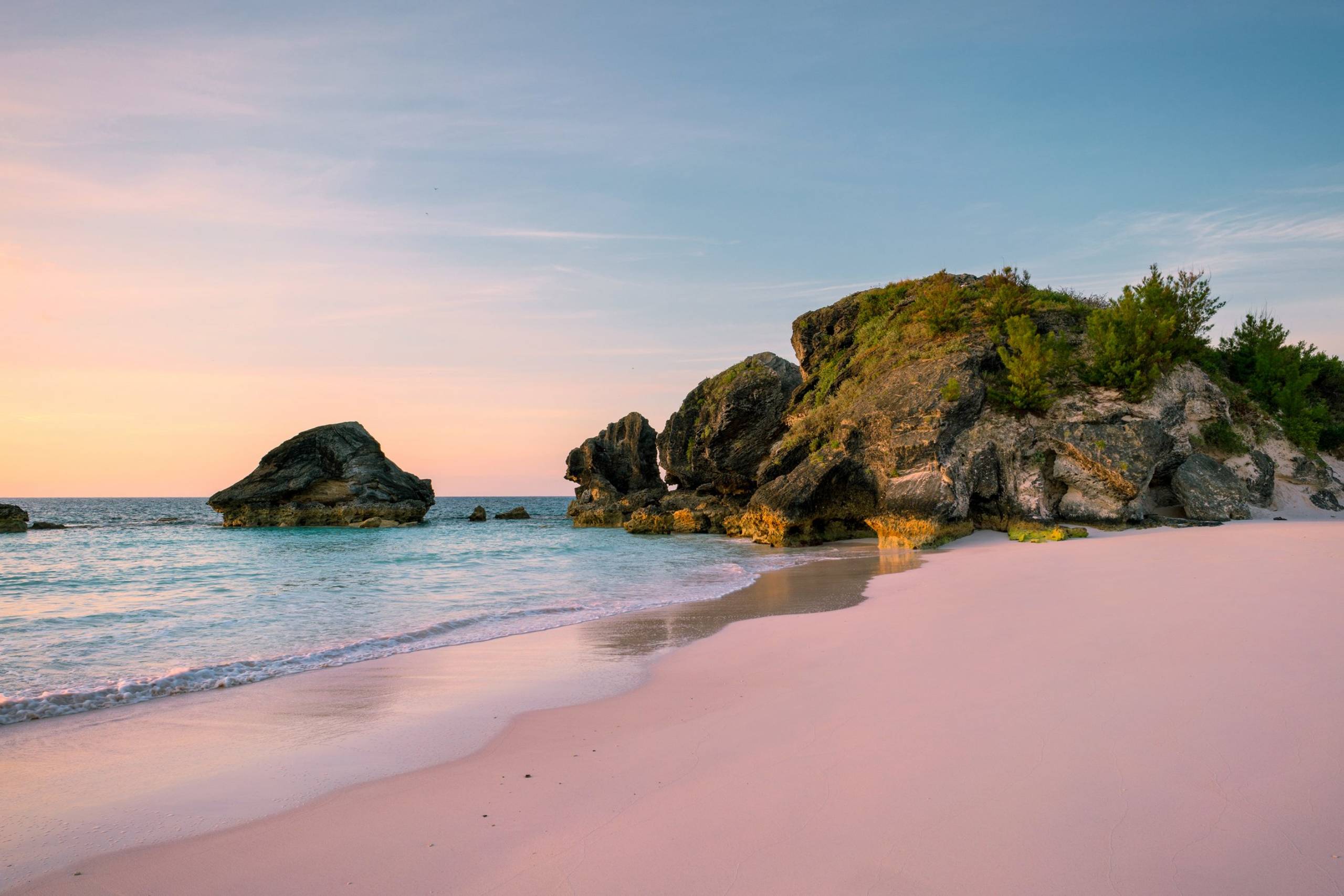Soft pink sand meets turquoise waters and dramatic rock formations under a pastel sky at sunrise.