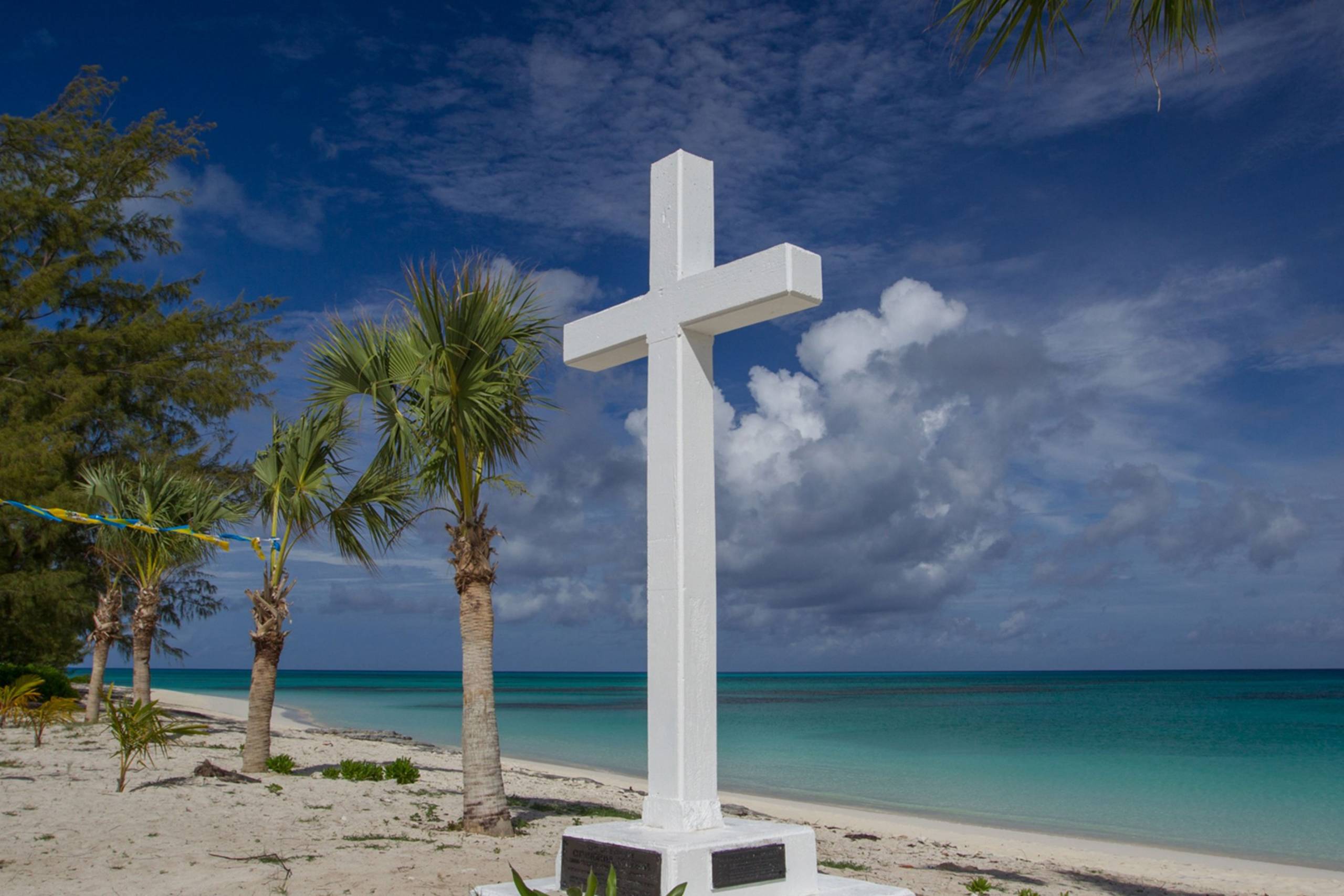A stark white cross stands sentinel on a sandy beach beside the vibrant turquoise waters of the Caribbean.