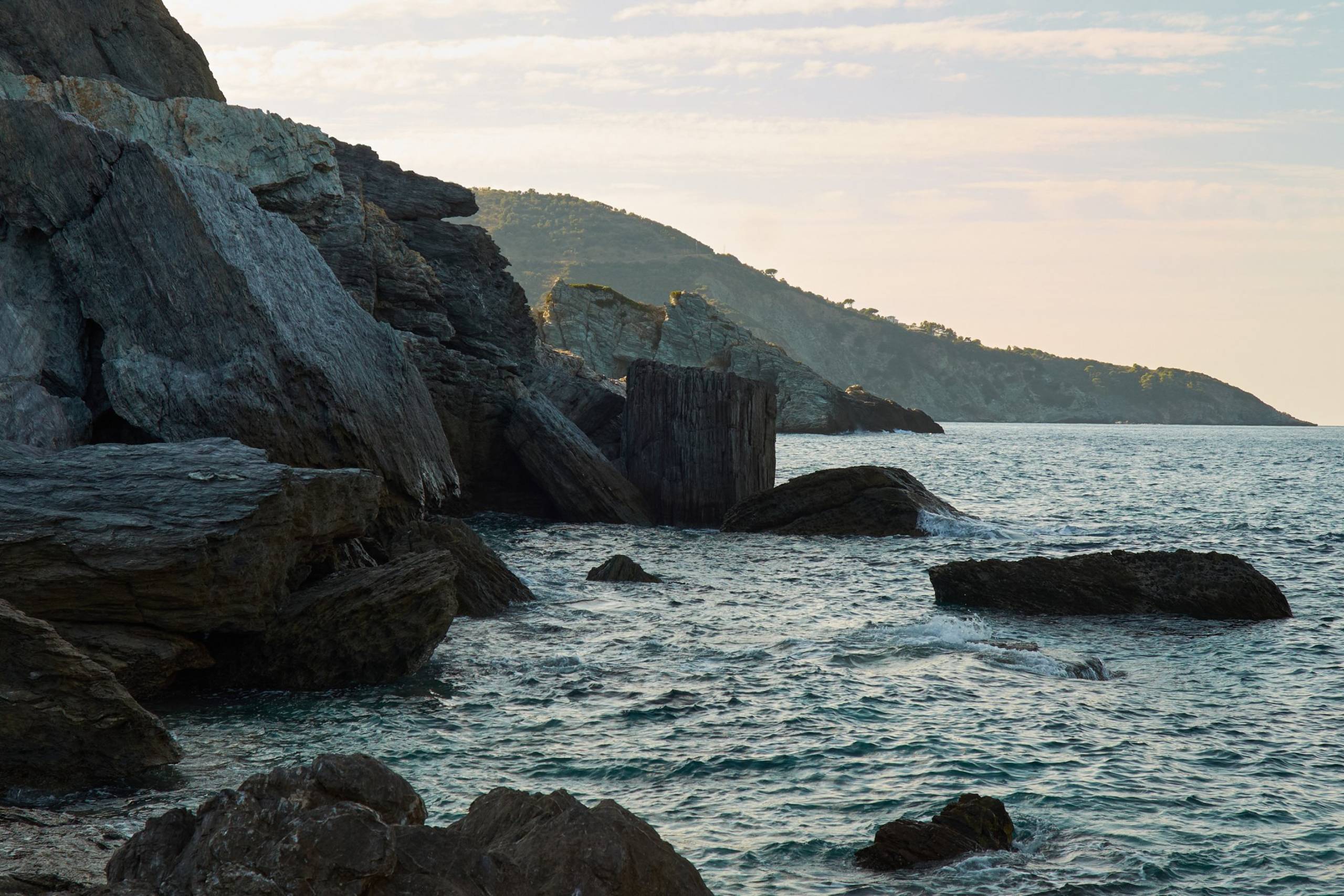 Rugged, ancient rocks meet the gentle waves of the sea under a soft, hazy sky.