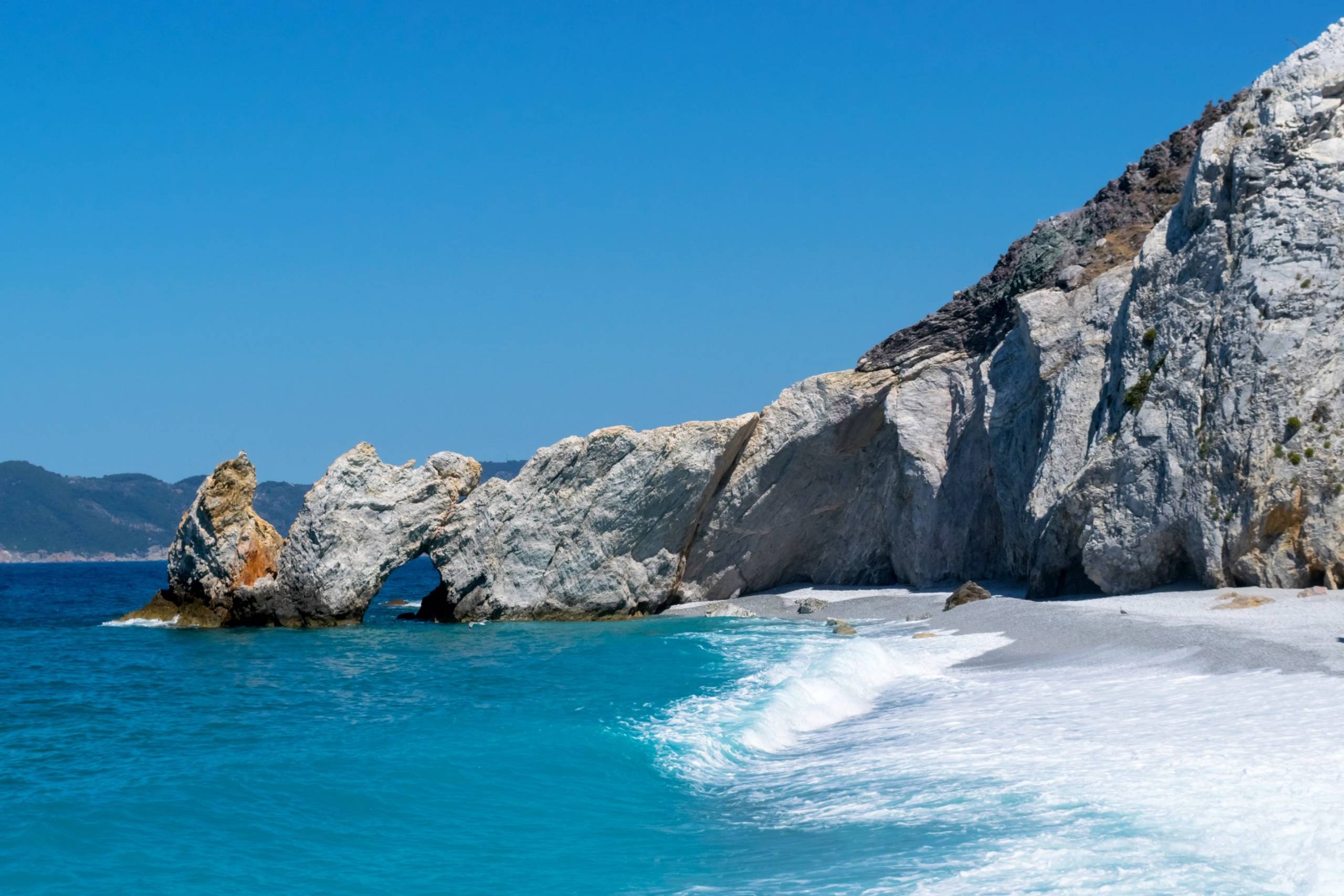 Turquoise waves crash onto a pebble beach beneath dramatic white cliffs and a natural rock arch.