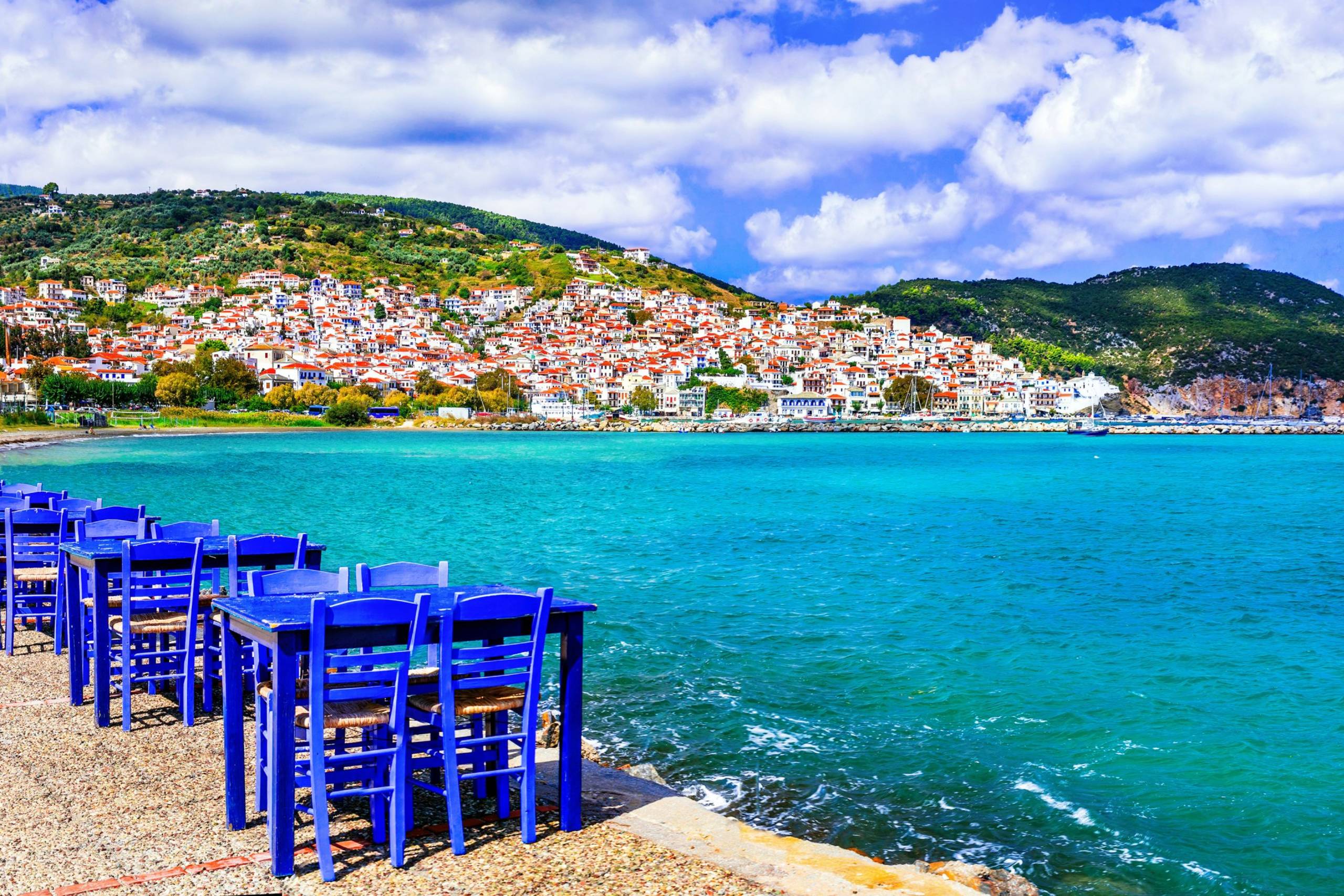 Blue tables and chairs await diners at a seaside cafe overlooking a charming Greek village nestled on a hillside.